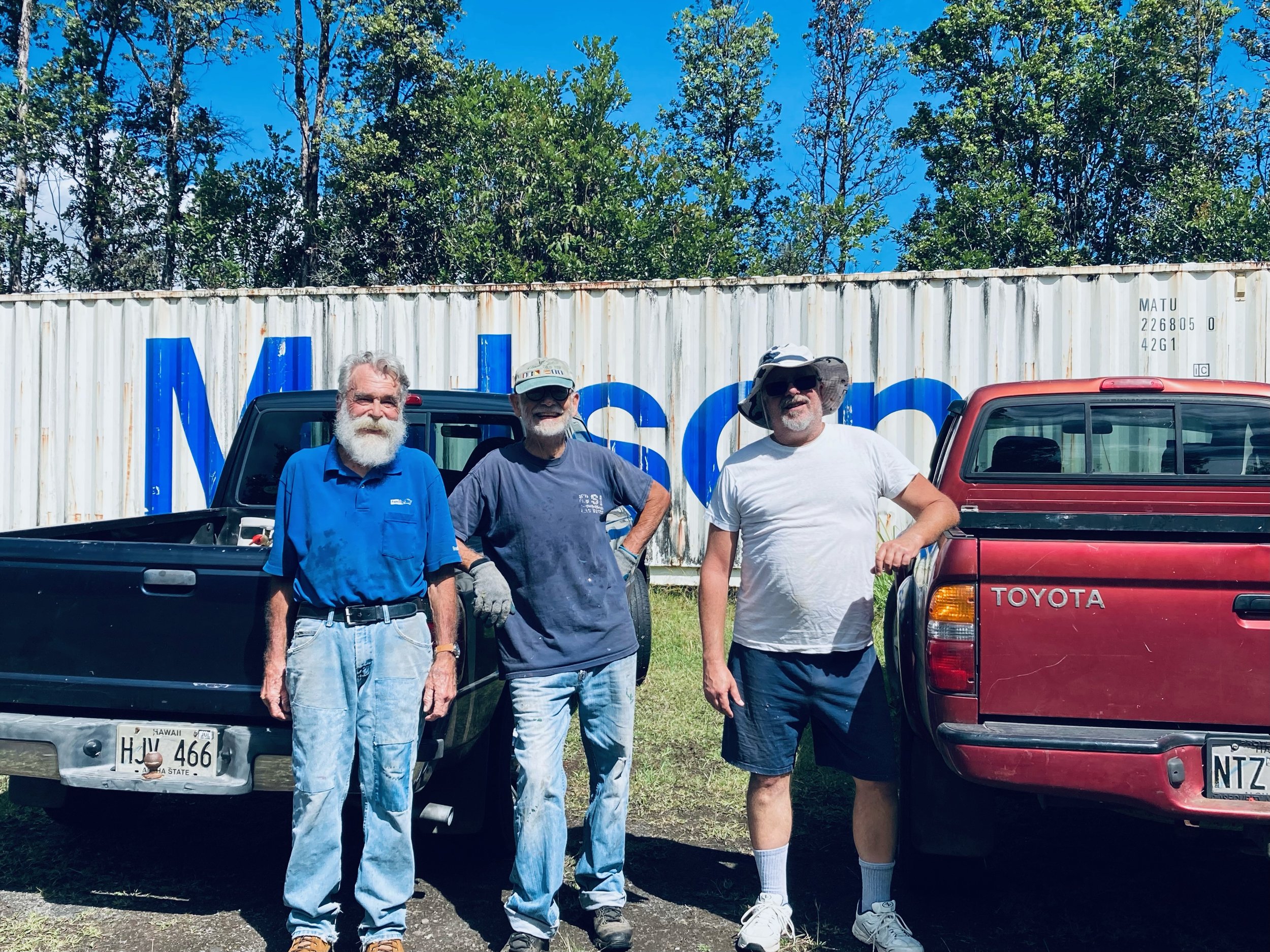 Three older men standing in front of a large shipping container and two pickup trucks, outdoors on a sunny day with trees and a blue sky in the background.