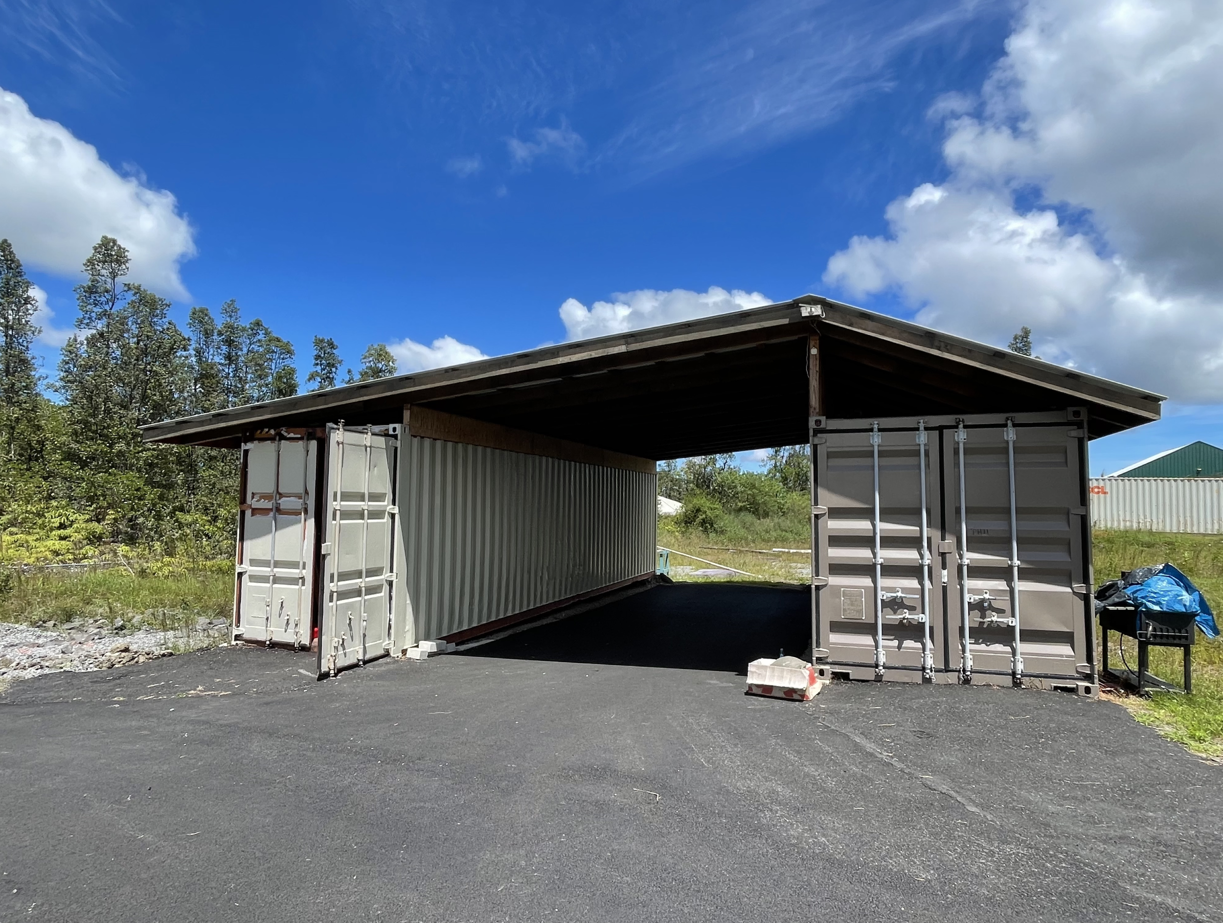 Two shipping containers are arranged to create a covered structure. One container with their doors open and the other with the doors closed. The structure is situated outdoors on a paved surface with trees and a blue sky with scattered clouds in the background.