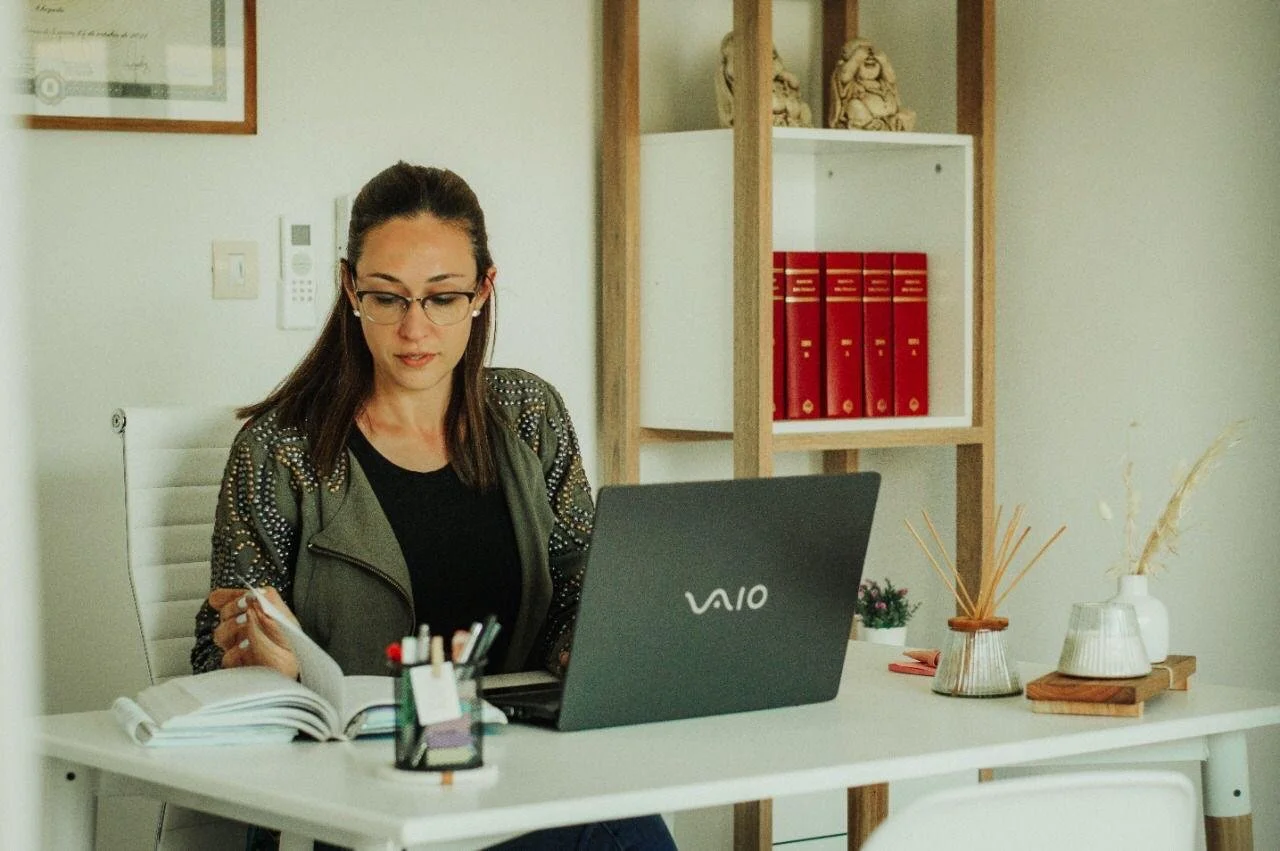 Mujer en una oficina moderna trabajando en su computadora portátil VAIO, revisando documentos con una libreta y bolígrafos, con estantería con carpetas rojas y decoraciones de madera en el fondo.