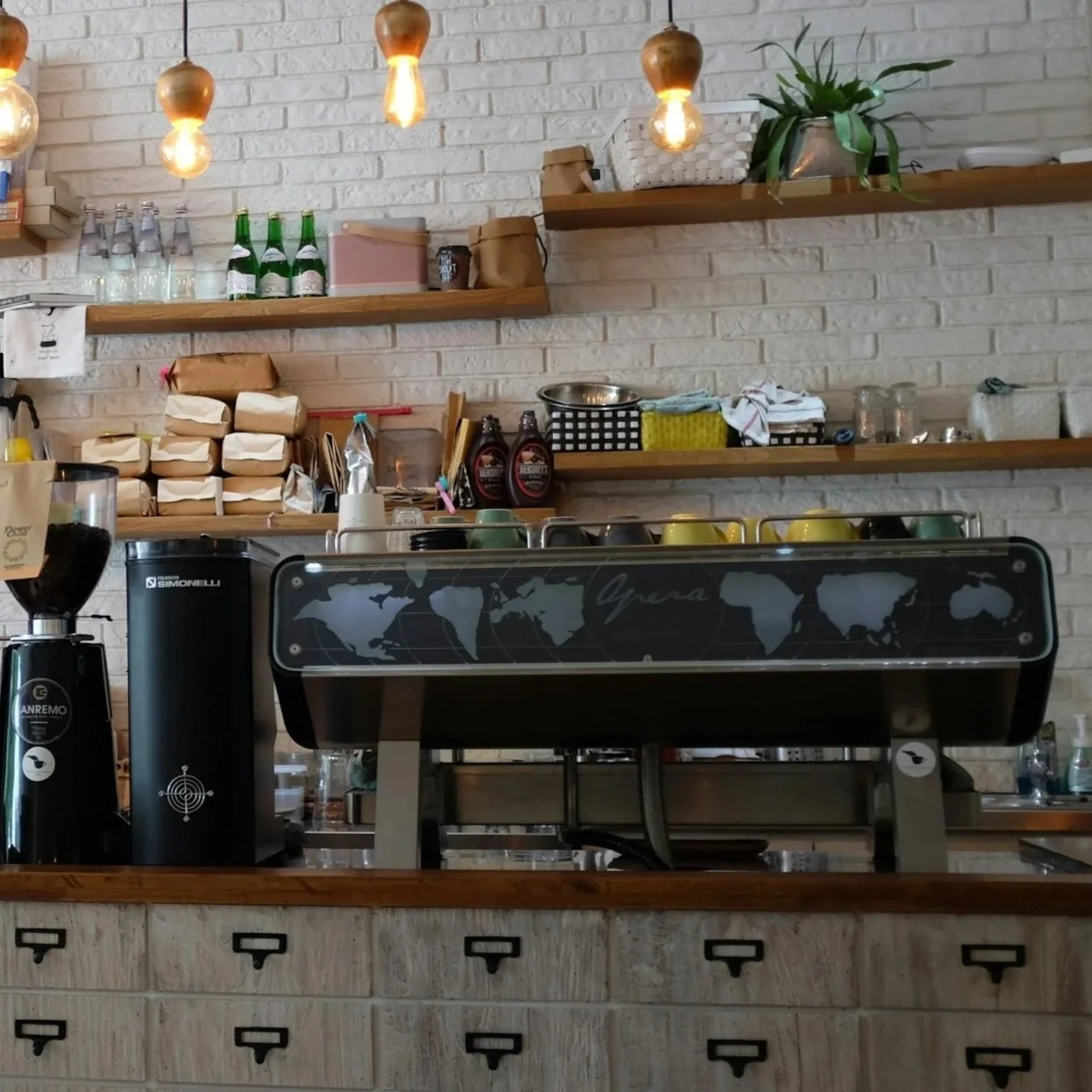 A coffee shop counter with an espresso machine, a grinder, and wooden shelves holding coffee supplies, bottles, and glassware. The background features a white brick wall with hanging lightbulbs, a plant, and various containers and packaging.