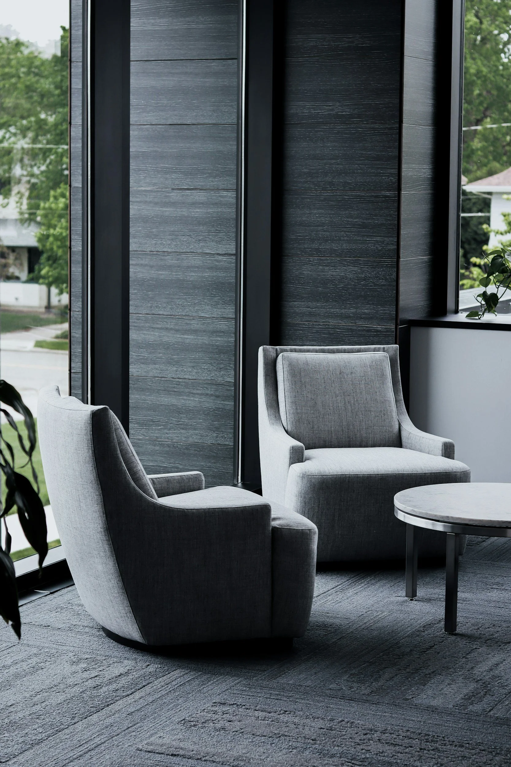 Modern interior with two gray upholstered armchairs and a round black table on a gray carpet. Large windows with black trim reveal green trees and residential houses outside.