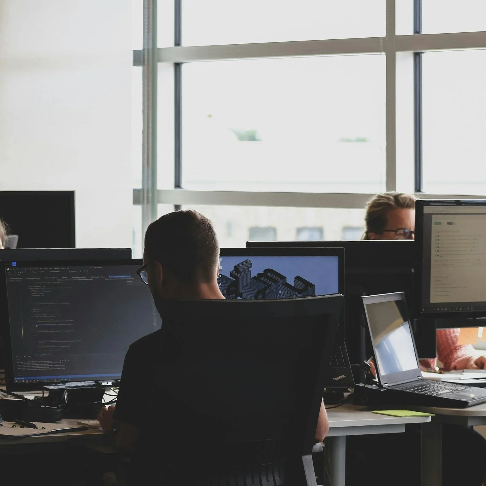 People working at computer desks in an office with large windows letting in natural light.