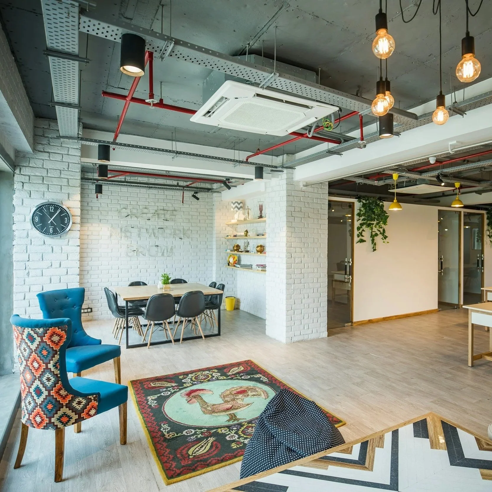 Modern interior space with a white brick wall, a round black wall clock, a wooden table with black chairs, a blue armchair with a colorful patterned cushion, a patterned rug, a bookshelf with decorative items, hanging pendant lights, exposed ceiling with red pipes, and a potted plant.