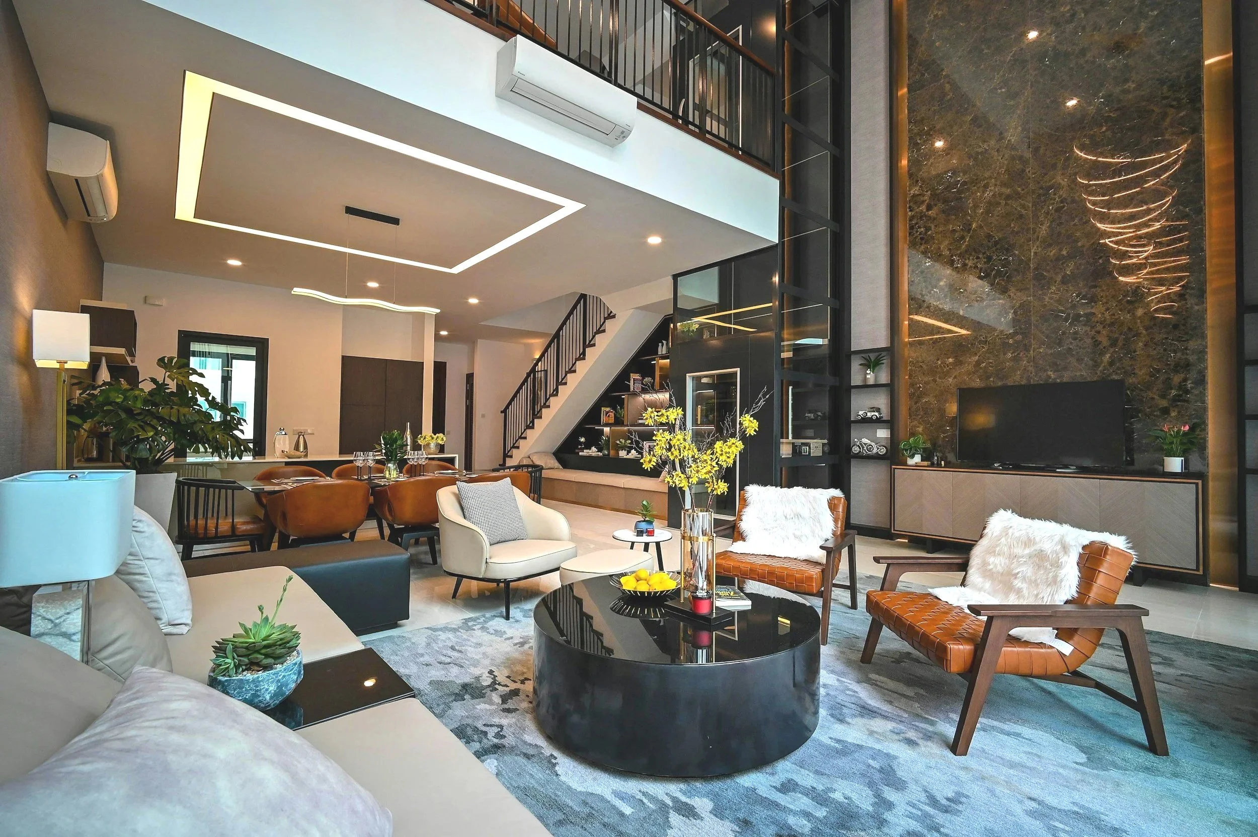 Interior view of a modern living room with beige and brown furniture, a black glossy round coffee table, decorative plants, and a staircase with a metal railing. The room has a high ceiling with recessed lighting, a marble accent wall, and a flat-screen TV. There is a mix of contemporary and cozy decor elements, including pillows and flowers.
