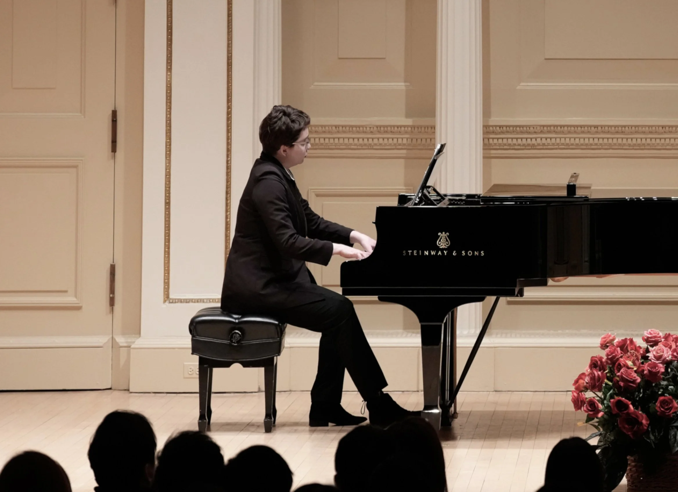 A person playing a grand piano on stage, wearing glasses and with short dark hair, dressed in a black suit, in a room with beige walls and decorative molding, audience members visible in the foreground.