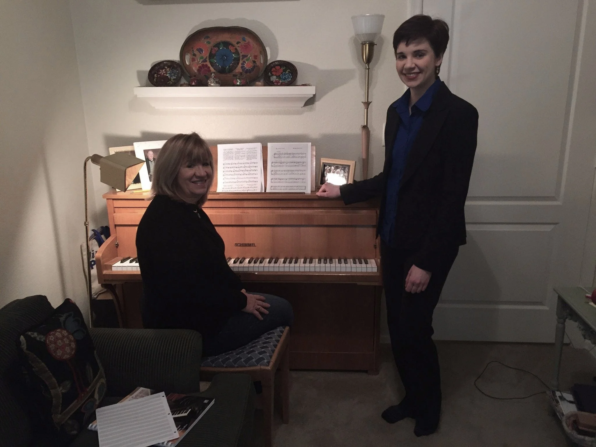 Two women in formal attire, one sitting at a piano and the other standing beside her, smiling in a home setting with decorative plates on a shelf in the background.