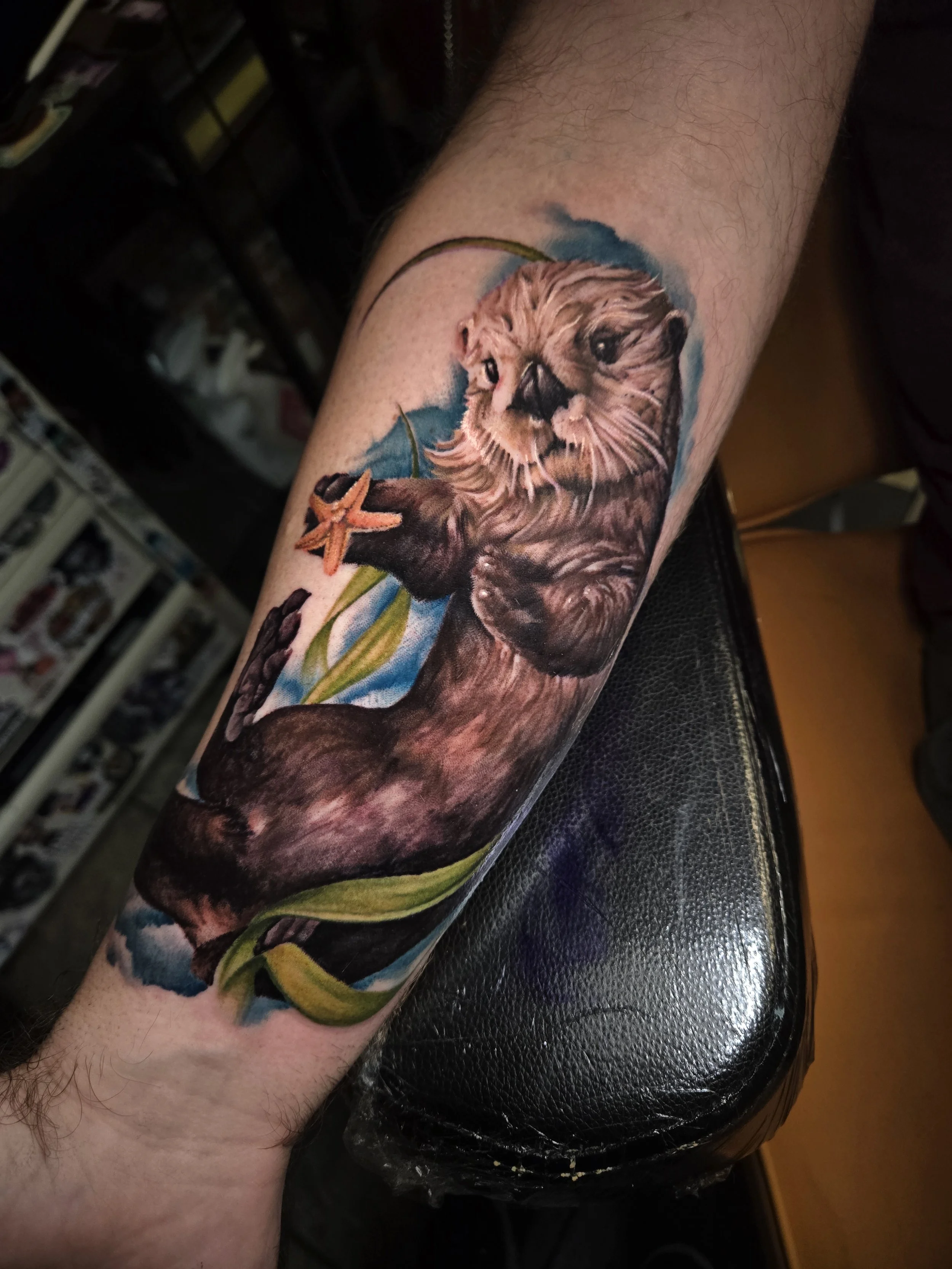 Colorful tattoo of an otter on a person's forearm, with blue and green backgrounds and an orange starfish near the paw.