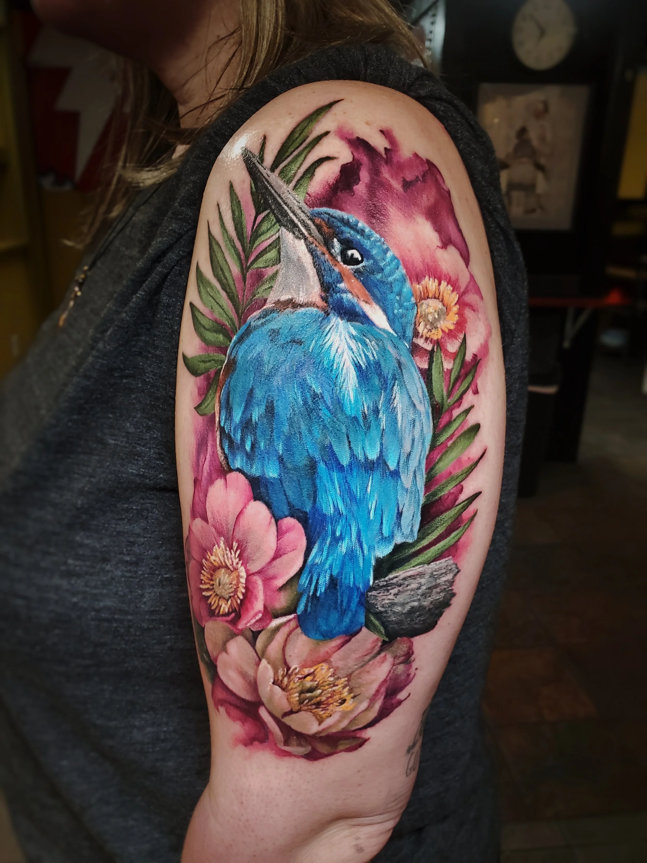 Colorful tattoo of a bluebird surrounded by pink flowers and green leaves on a person's shoulder.