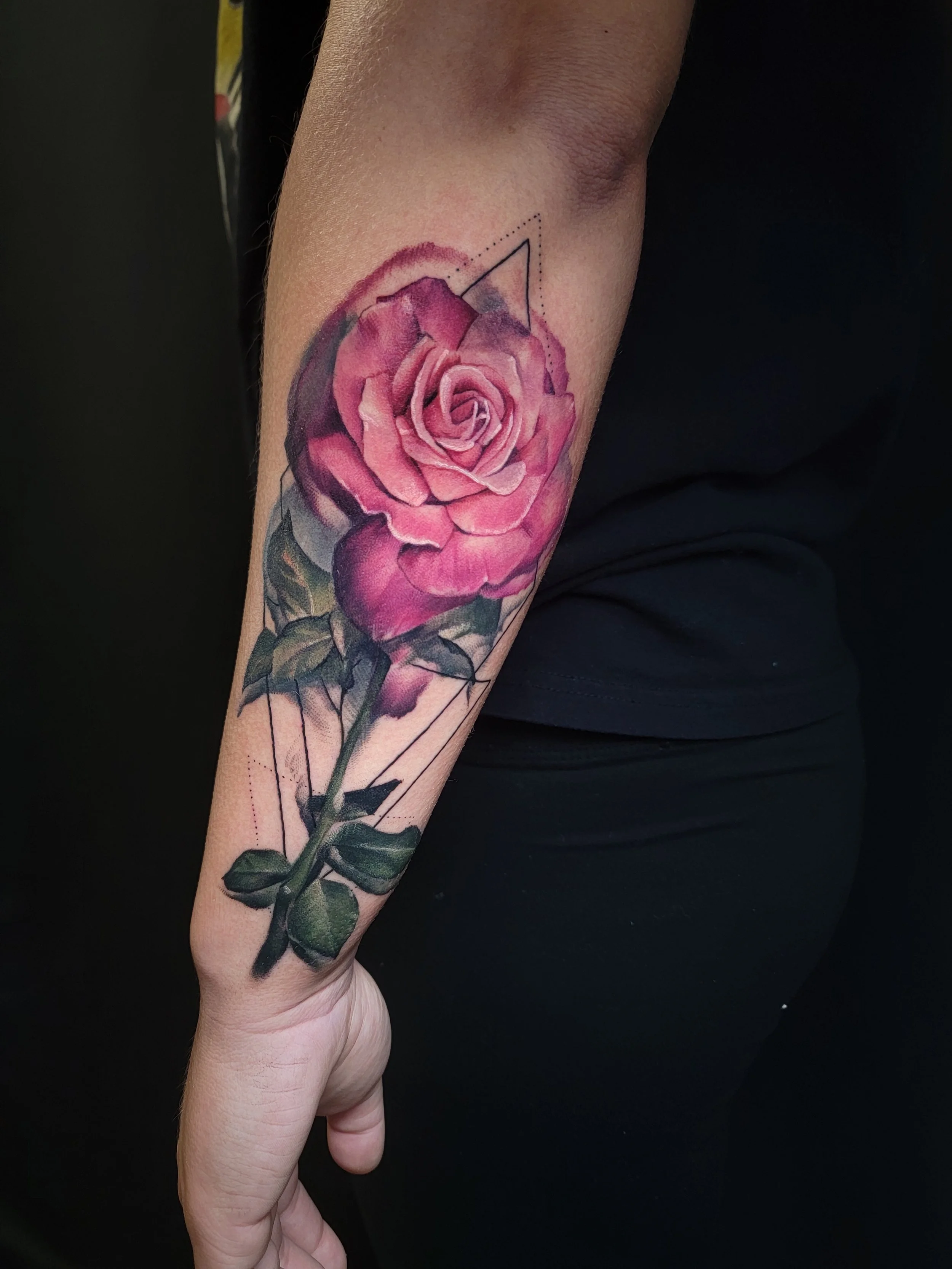 A person's forearm with a detailed, realistic pink and purple rose tattoo, including green leaves, with geometric line accents above the flower.