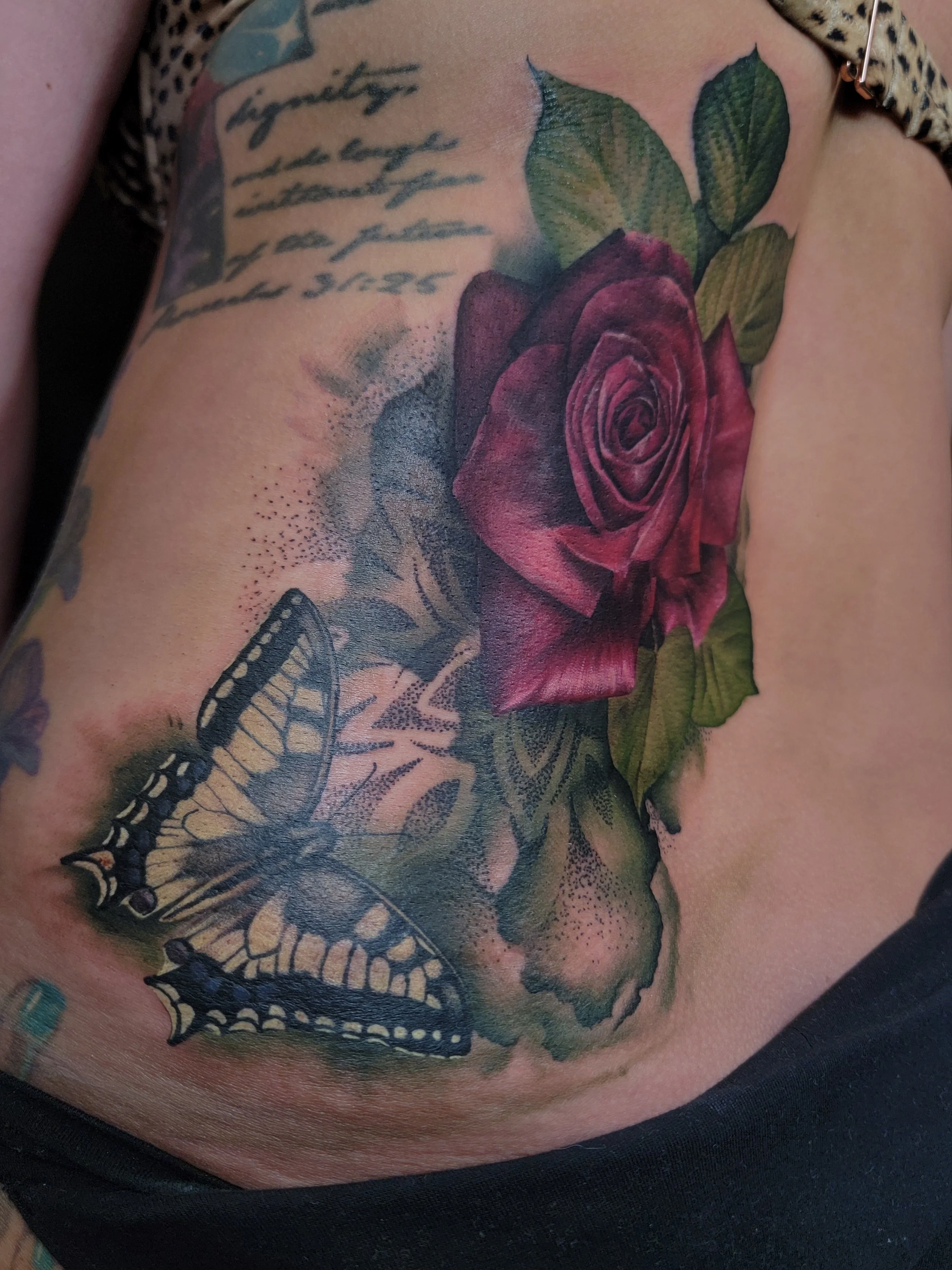 A tattoo of a red rose with green leaves, a butterfly, and cursive writing on the person's skin.