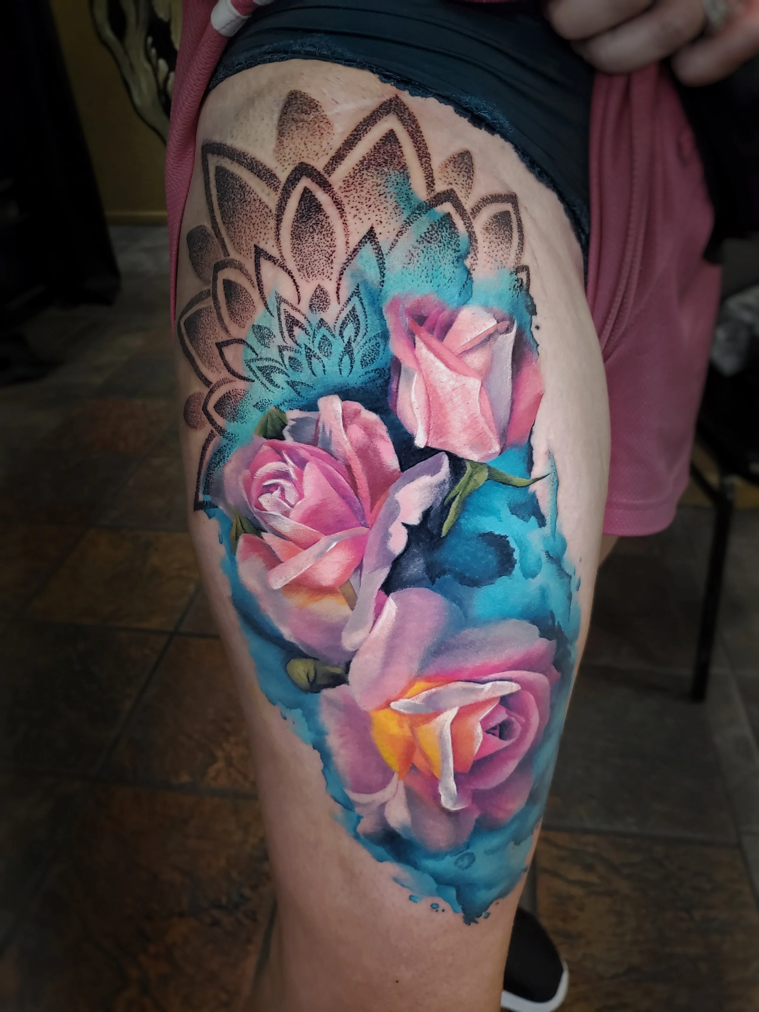 Tattoo of pink roses with green leaves and blue watercolor background on person's upper leg.