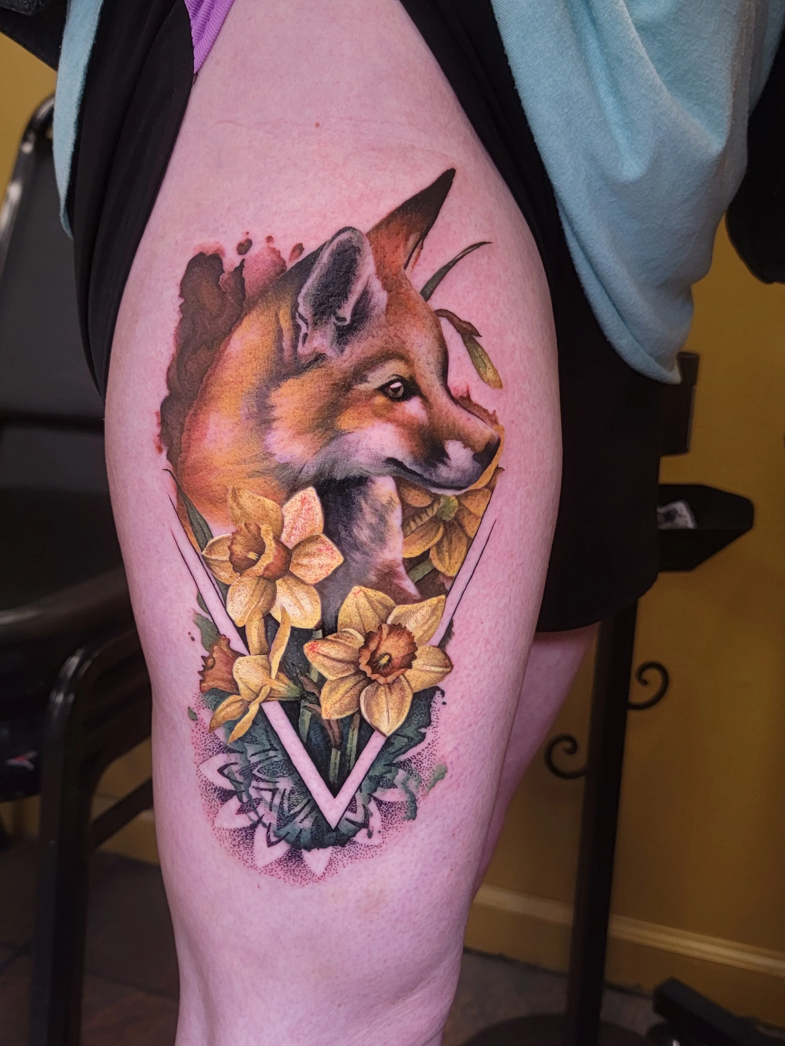 Tattoo of a fox surrounded by yellow daffodil flowers and green leaves, with a decorative border underneath.
