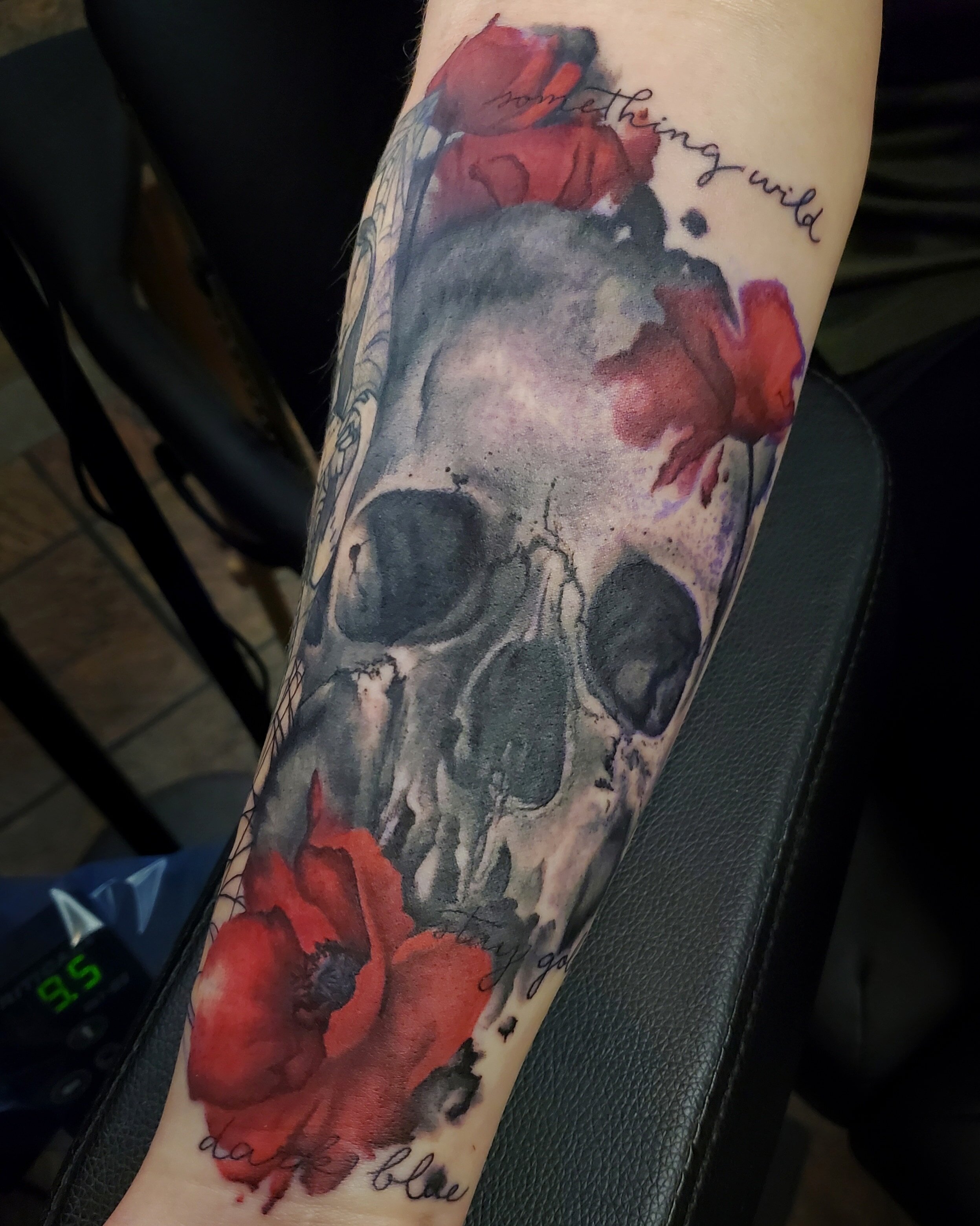 A tattoo of a realistic red and black flower, possibly a rose, with handwritten text above it that reads "honoring your wild" and below that "because I believe".