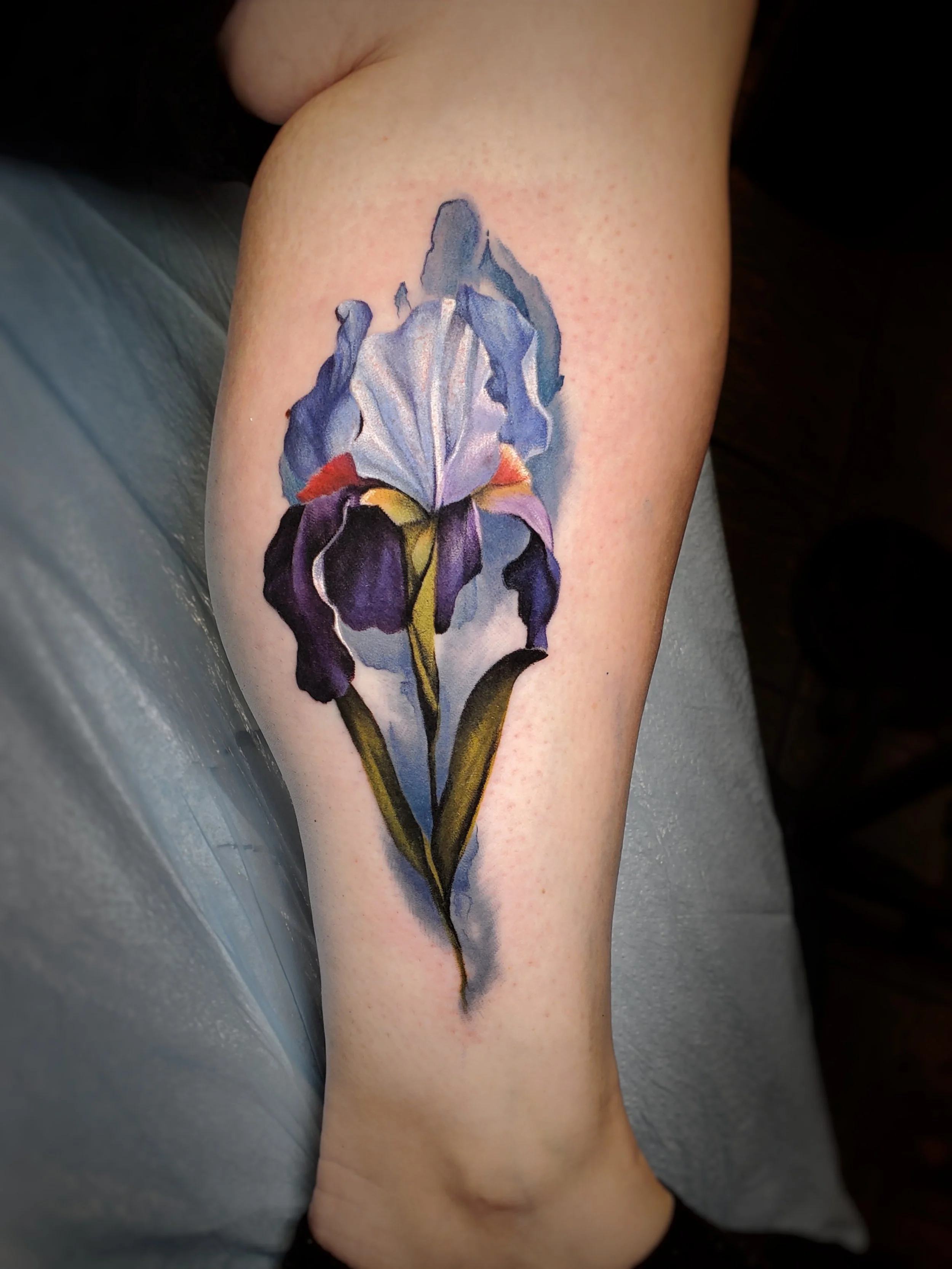 A realistic watercolor style tattoo of a purple iris flower on a person's calf