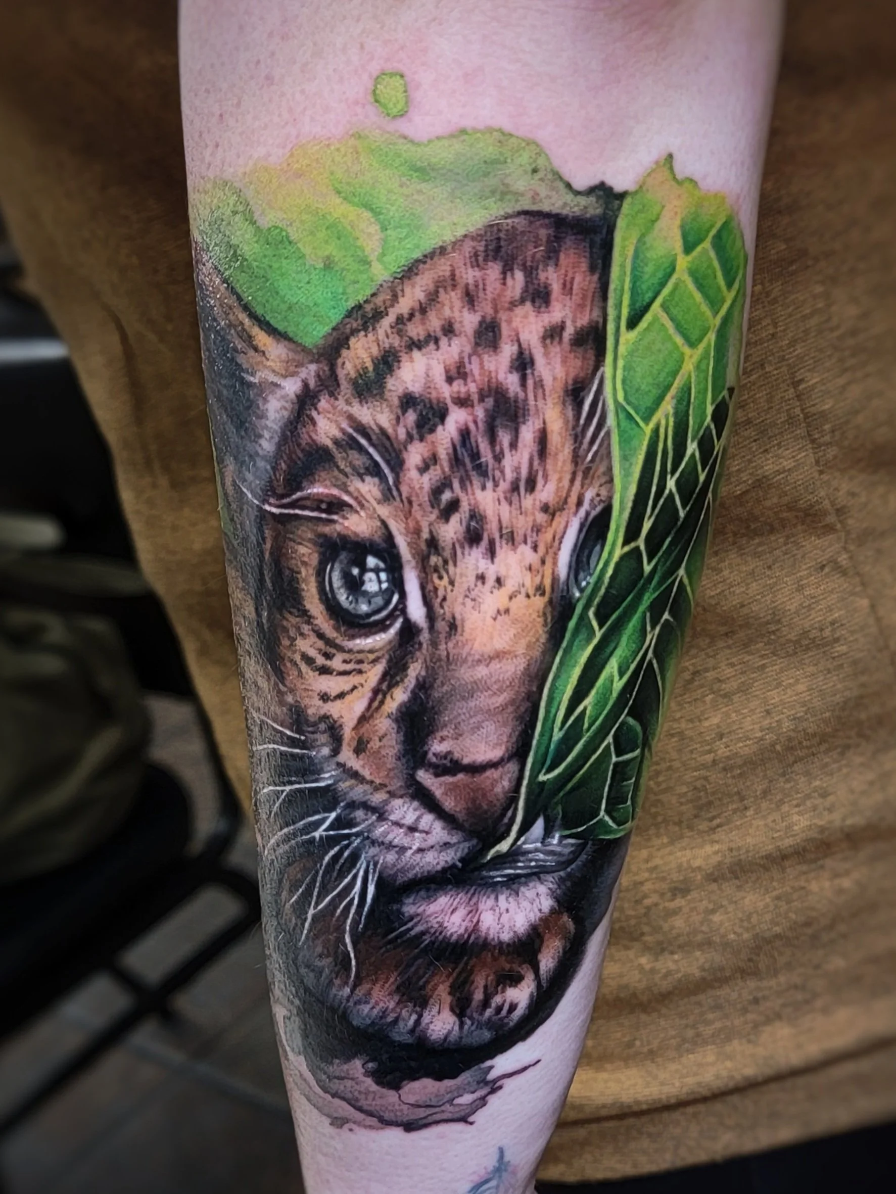 Tattoo of a kitten with detailed eyes, nose, whiskers, and fur, surrounded by green leaves.