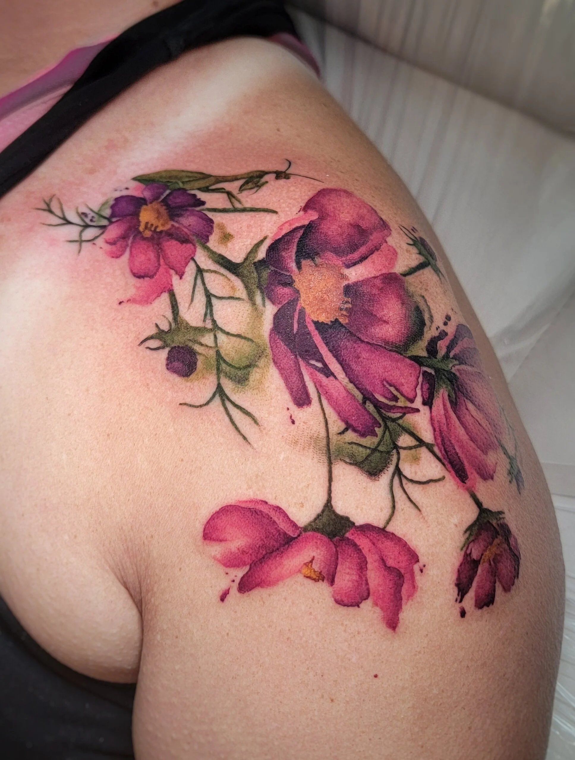 Close-up of a shoulder tattoo featuring vibrant pink and purple flowers with green stems and leaves.