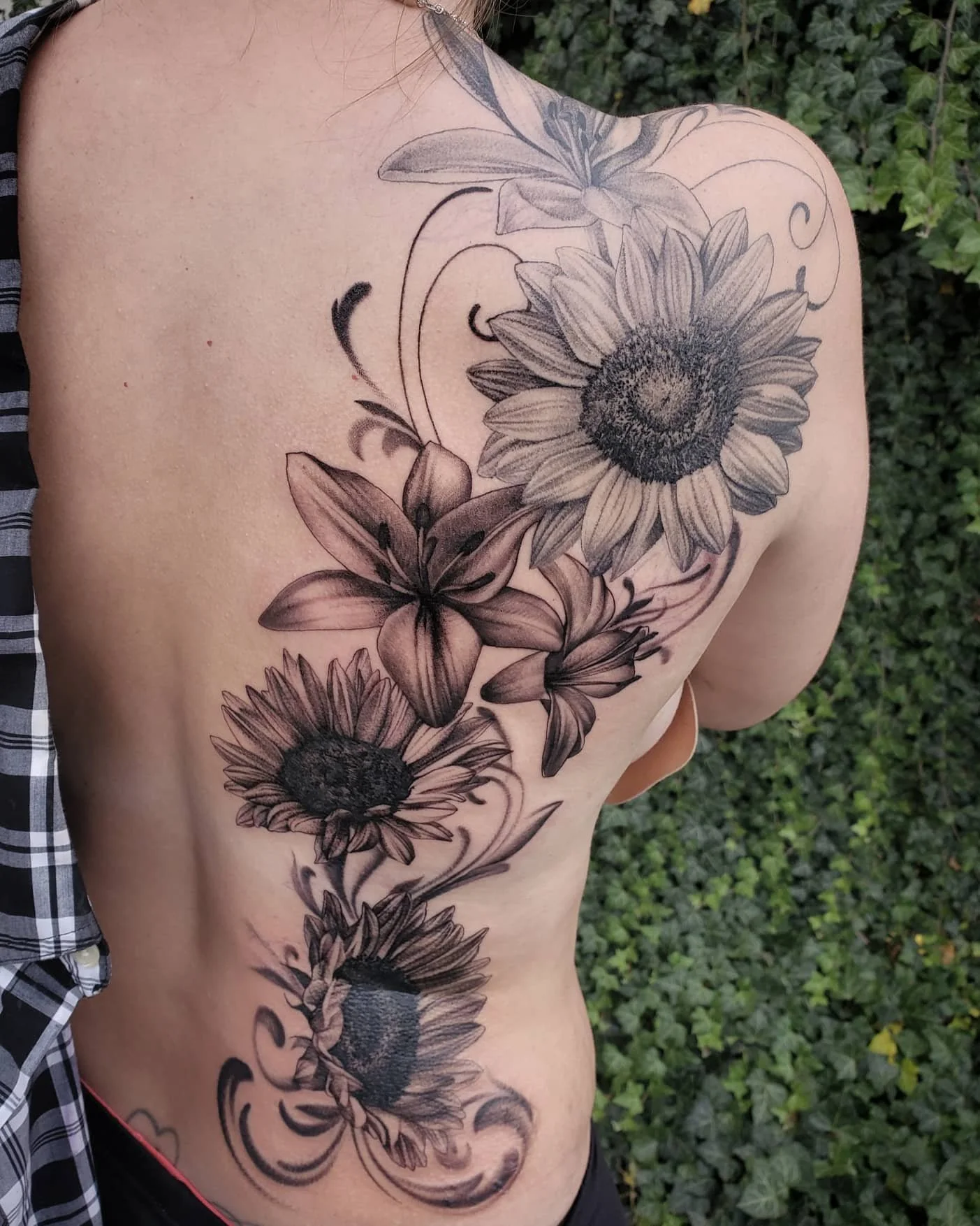 Black and white floral tattoo on person's left shoulder and side, featuring sunflowers, lilies, and decorative swirl designs, with green foliage background.