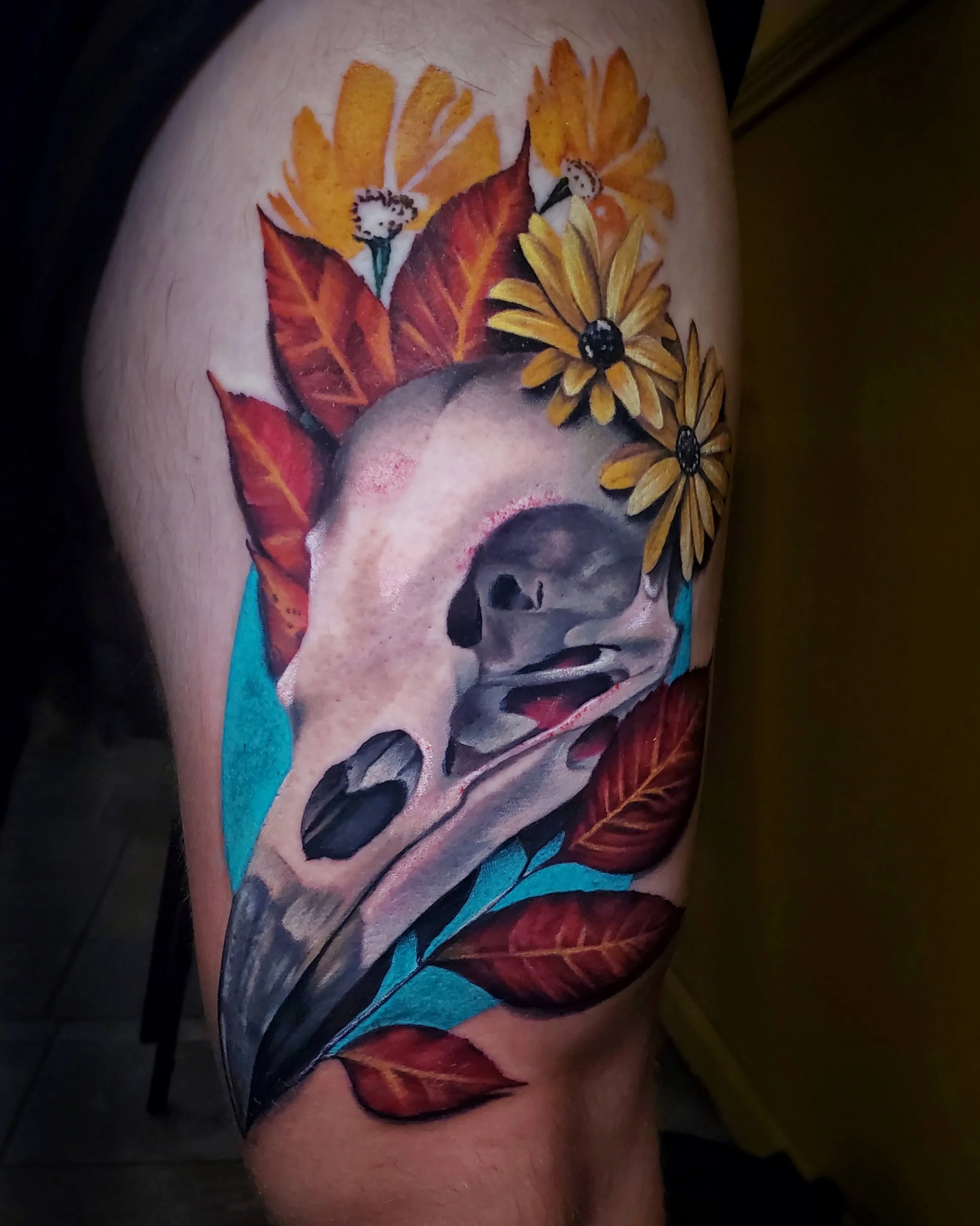 A tattoo on a person's thigh featuring colorful flowers, leaves, and a bird skull.