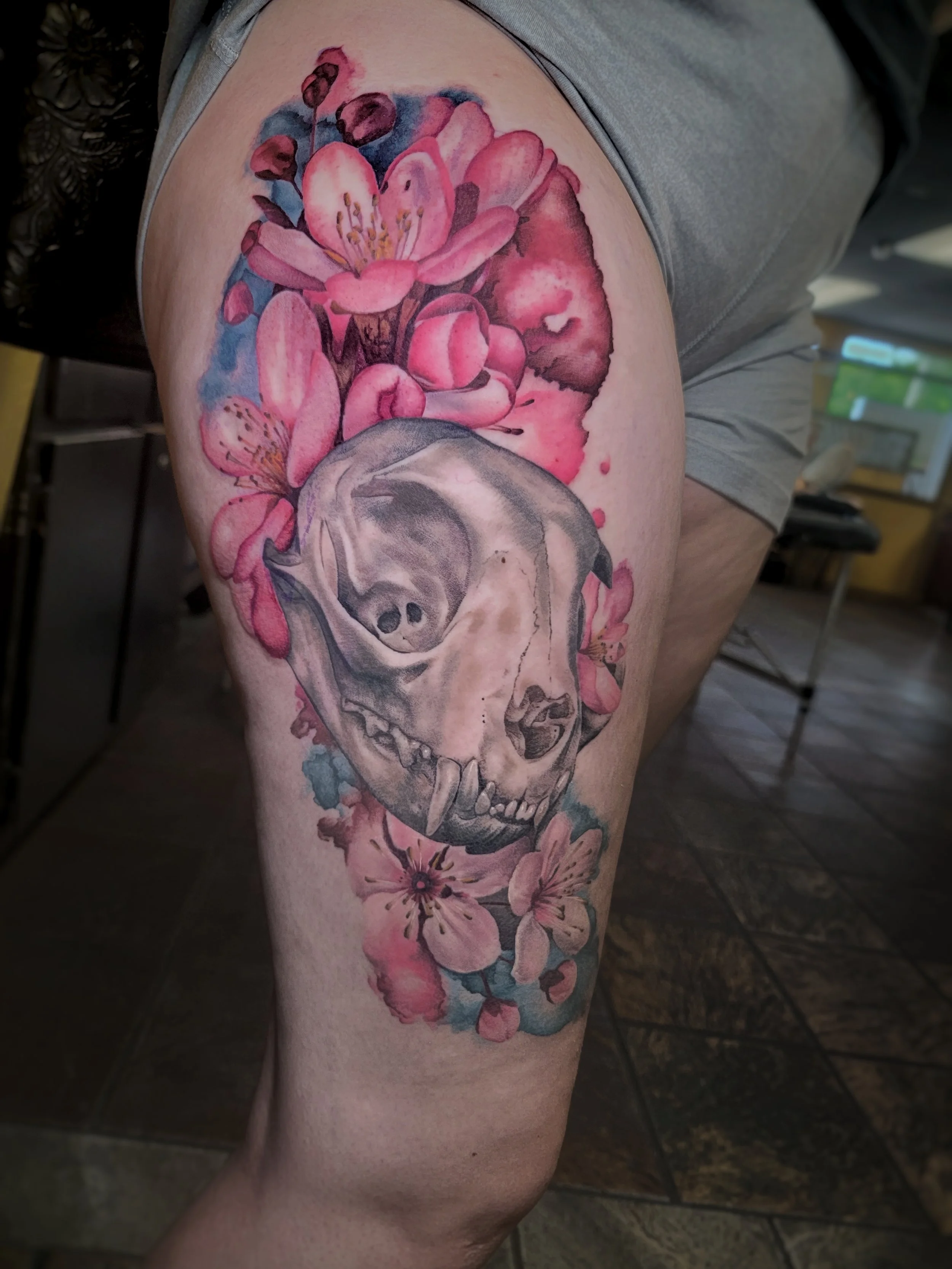 A tattoo of pink and red flowers with a cat skull below them on a person's upper thigh.