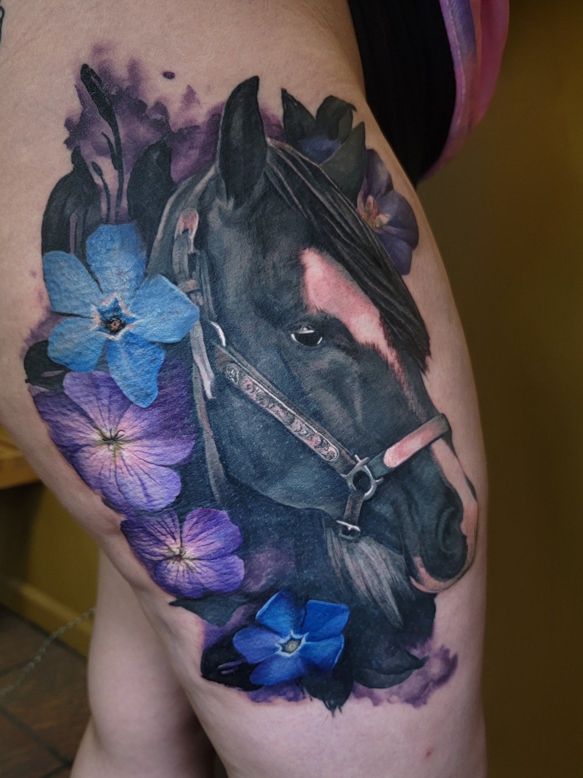 Tattoo of a black horse surrounded by purple and blue flowers on a person's upper thigh.