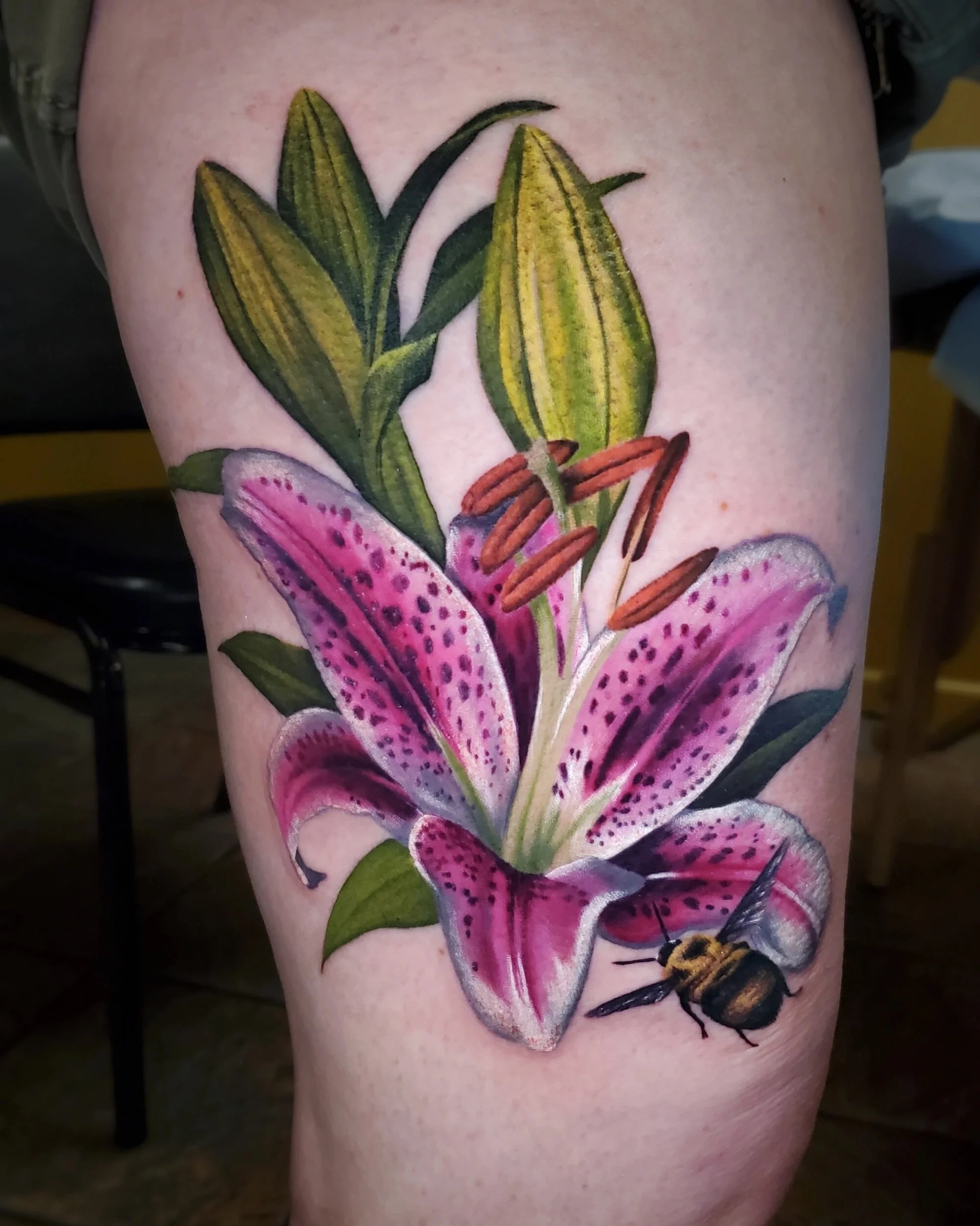 Tattoo of a pink lily flower with green leaves and orange-streaked pollen on a person's skin, featuring a bee near the flower.