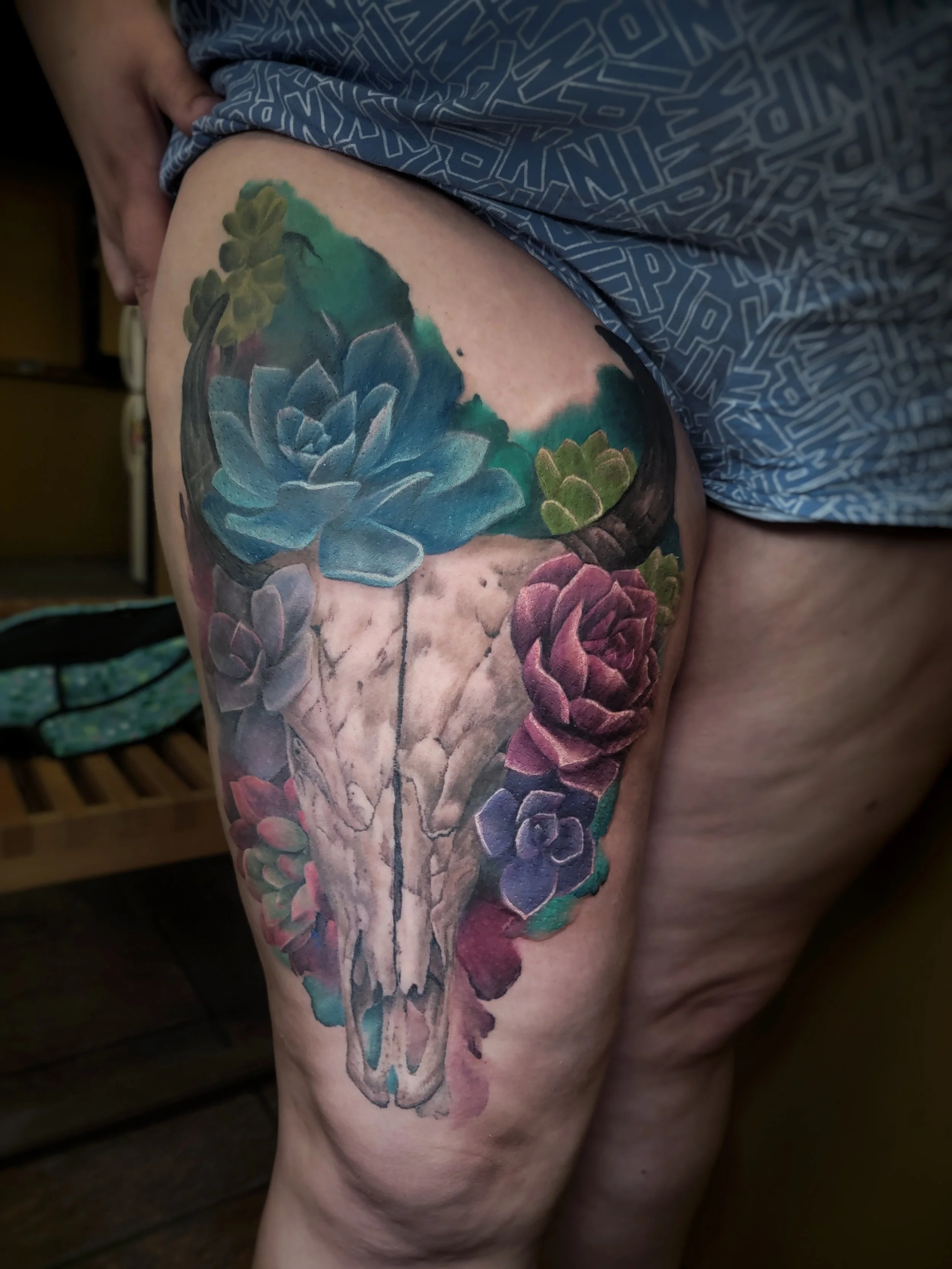 Colorful tattoo of succulents surrounding a bull skull on a person's thigh.