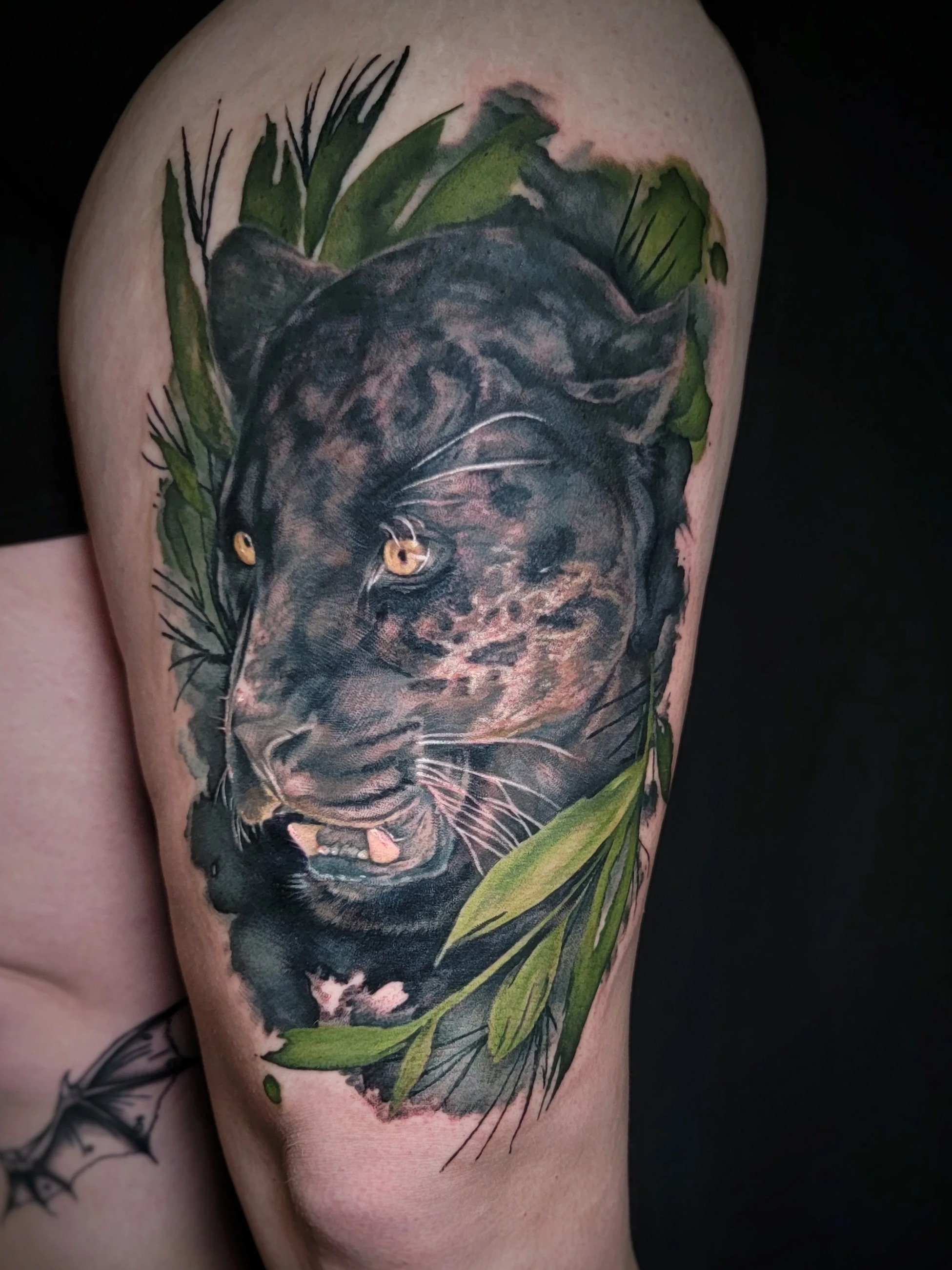 A tattoo of a black panther's face surrounded by green leaves on a person's thigh