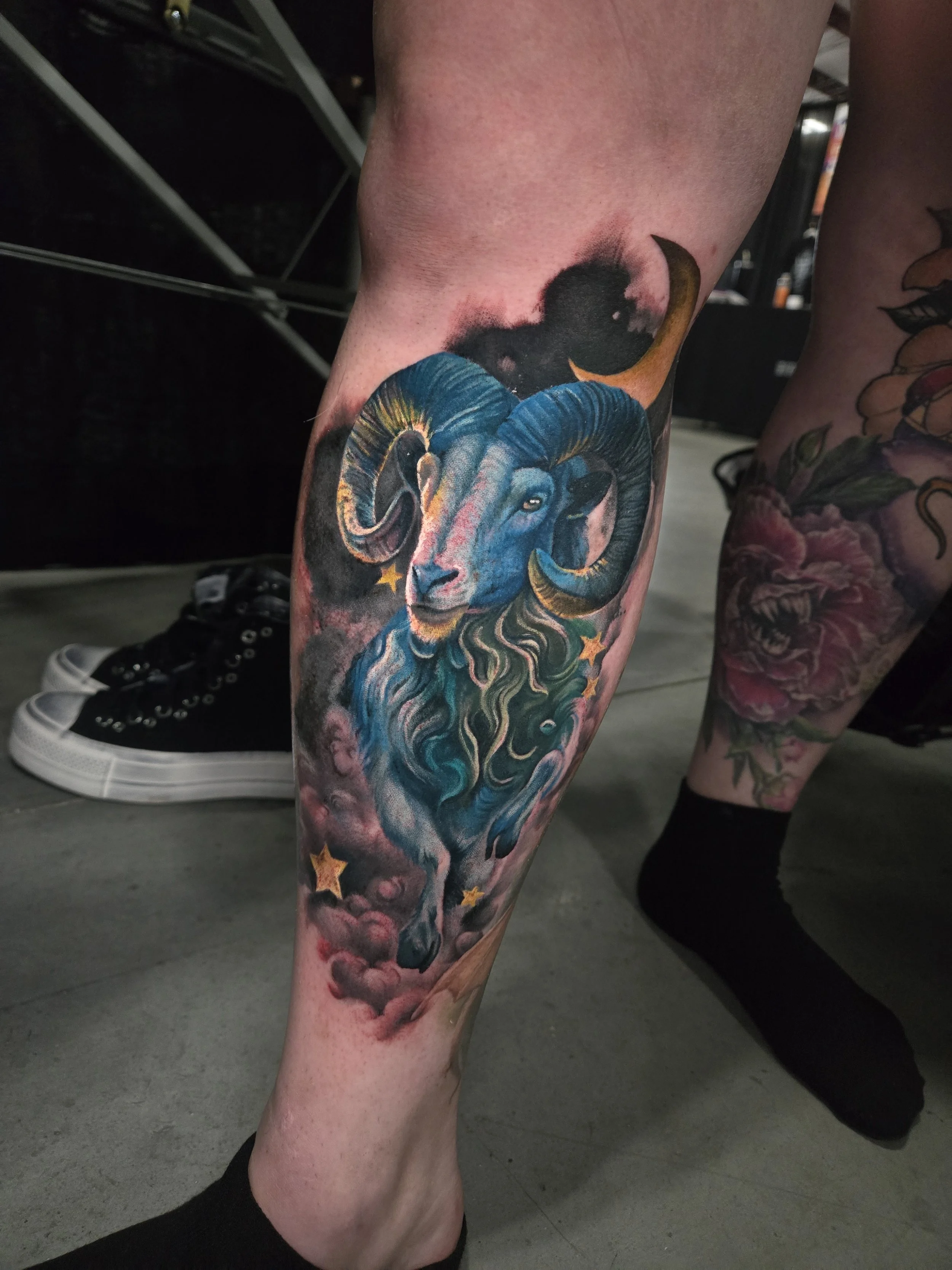Tattoo of a ram's head with big curled horns on a person's leg, surrounded by colorful elements and stars.