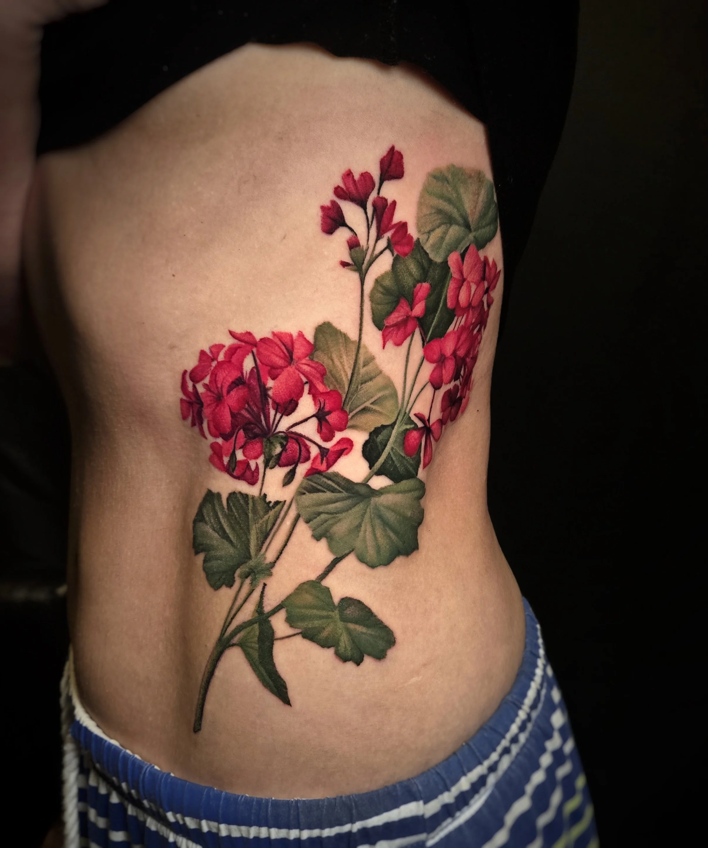 A close-up of a person's rib tattoo featuring a detailed tattoo of a red flowering plant with green leaves.