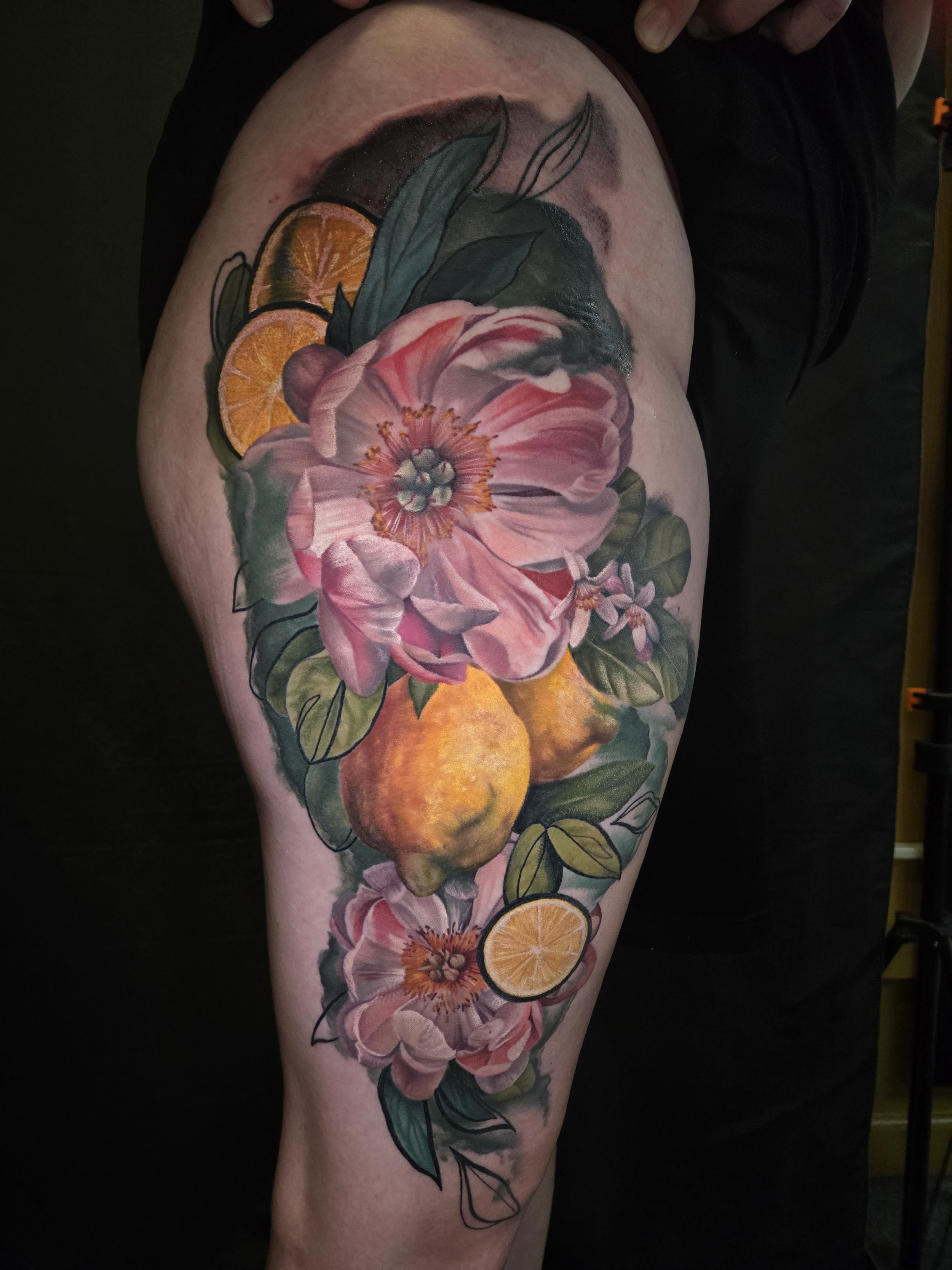 A tattoo on a person's thigh featuring pink and white flowers, green leaves, and slices of lemons