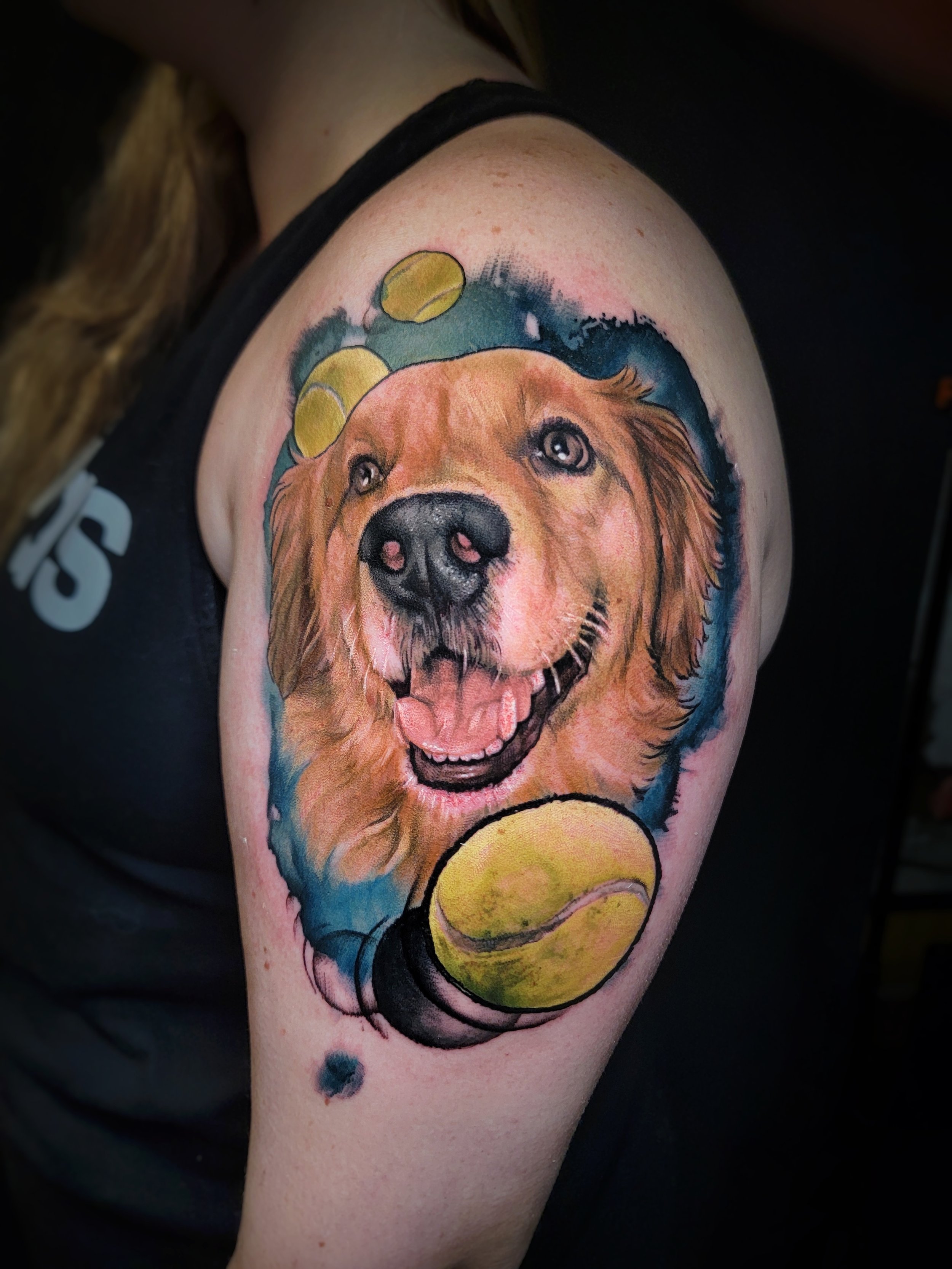 Colorful tattoo of a happy dog portrait with its tongue out, surrounded by yellow tennis balls, on the upper arm.
