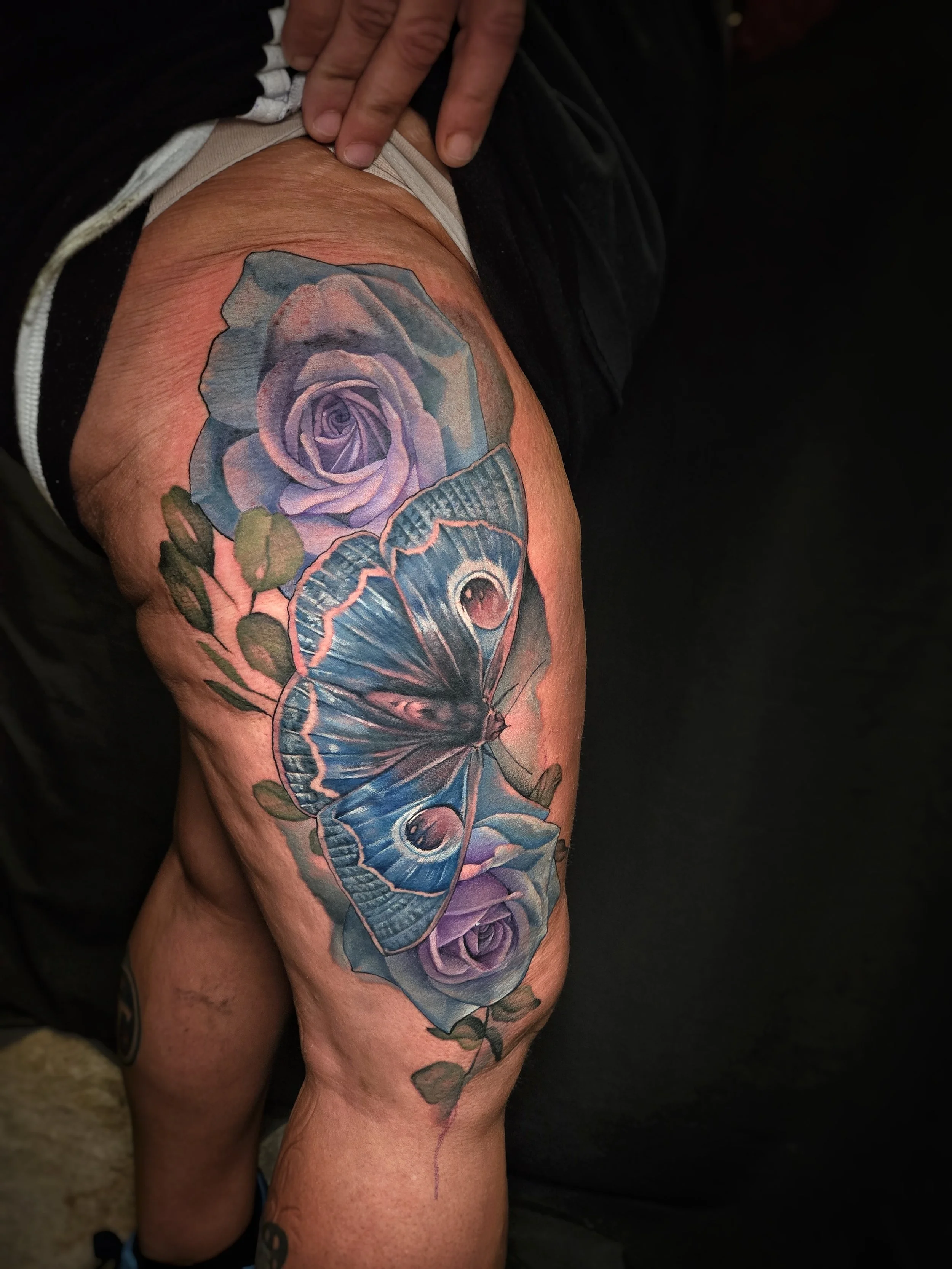 A person's arm with a colorful tattoo of a purple rose and a blue moth on the upper thigh