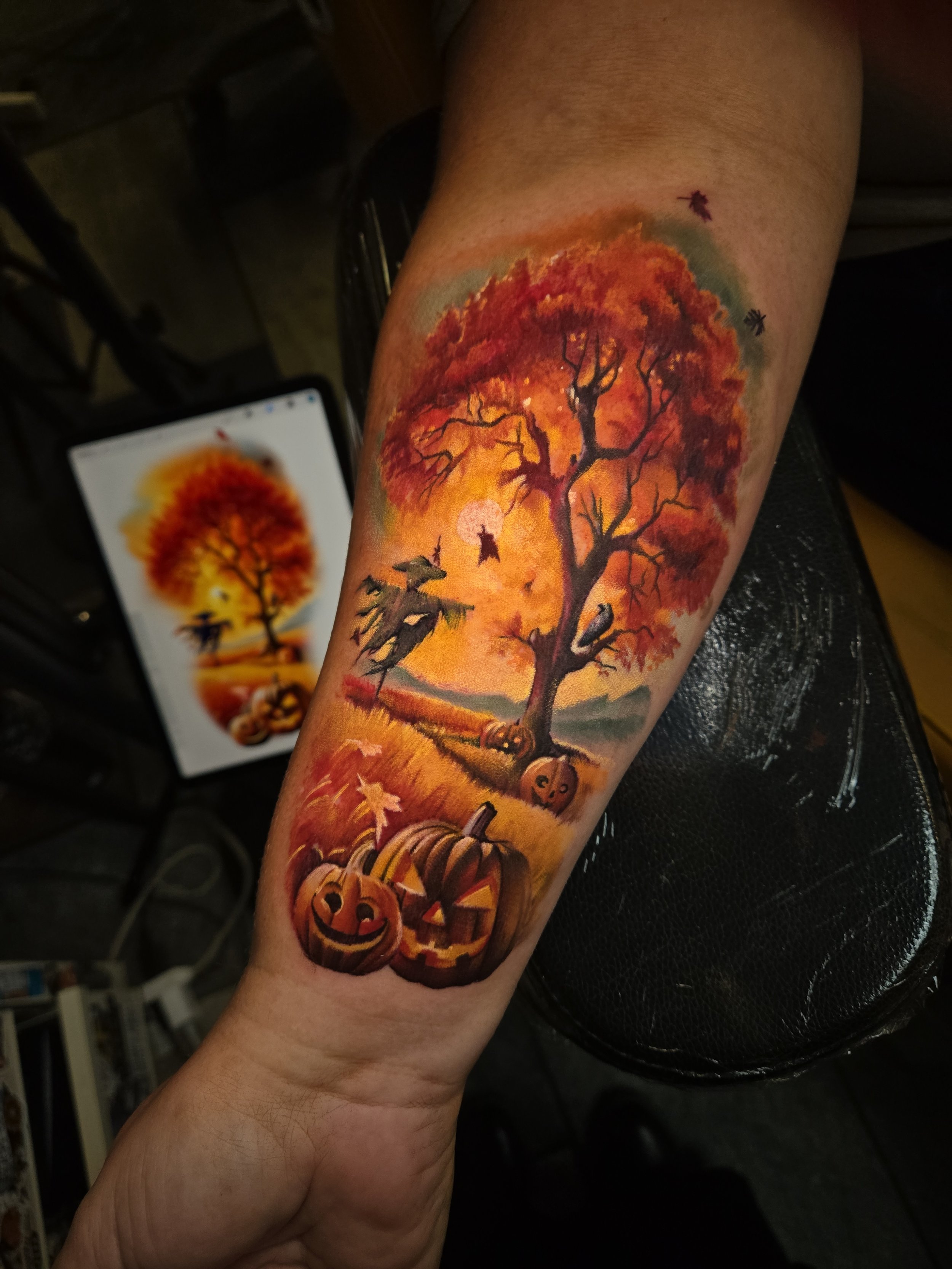 Colorful tattoo of a large autumn tree with red and orange leaves on a person's forearm, with Halloween-themed pumpkins and fall scenery in the background.