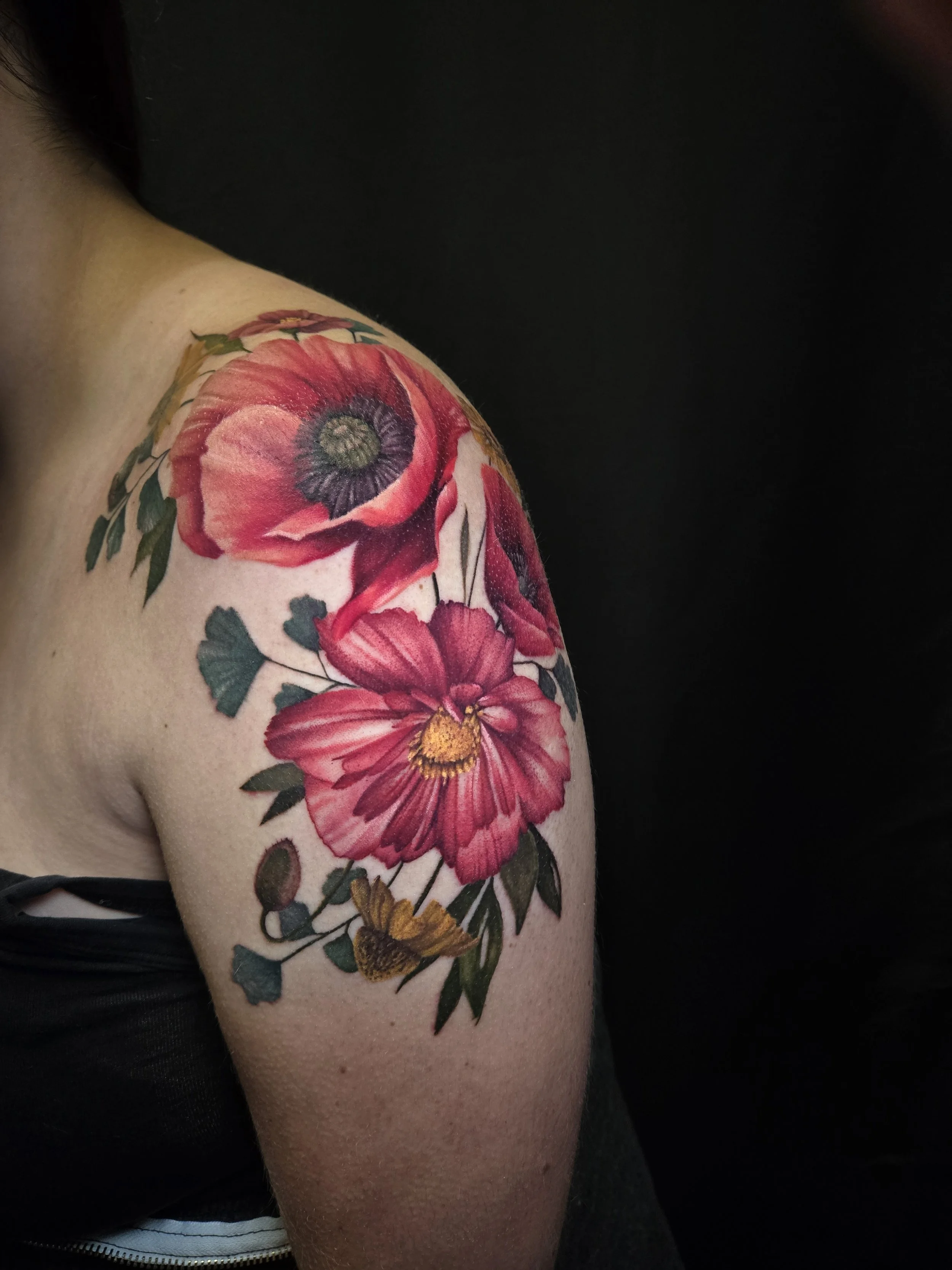 A person with a large colorful floral tattoo on their shoulder and upper arm, featuring red, pink, and yellow flowers with green leaves, against a black background.