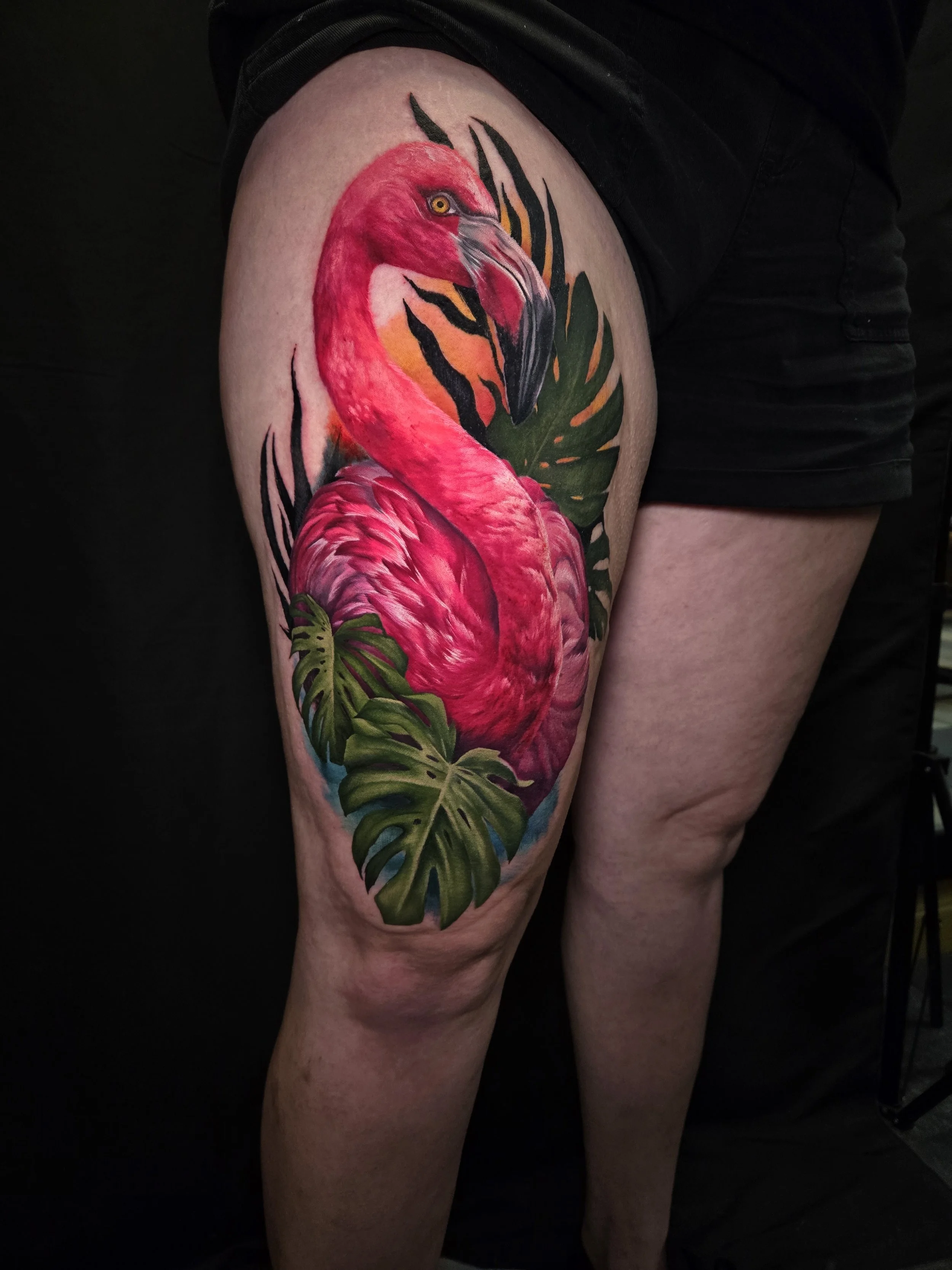 Colorful tattoo of a pink flamingo with a black beak and yellow eye, surrounded by green tropical leaves, on a person's thigh.
