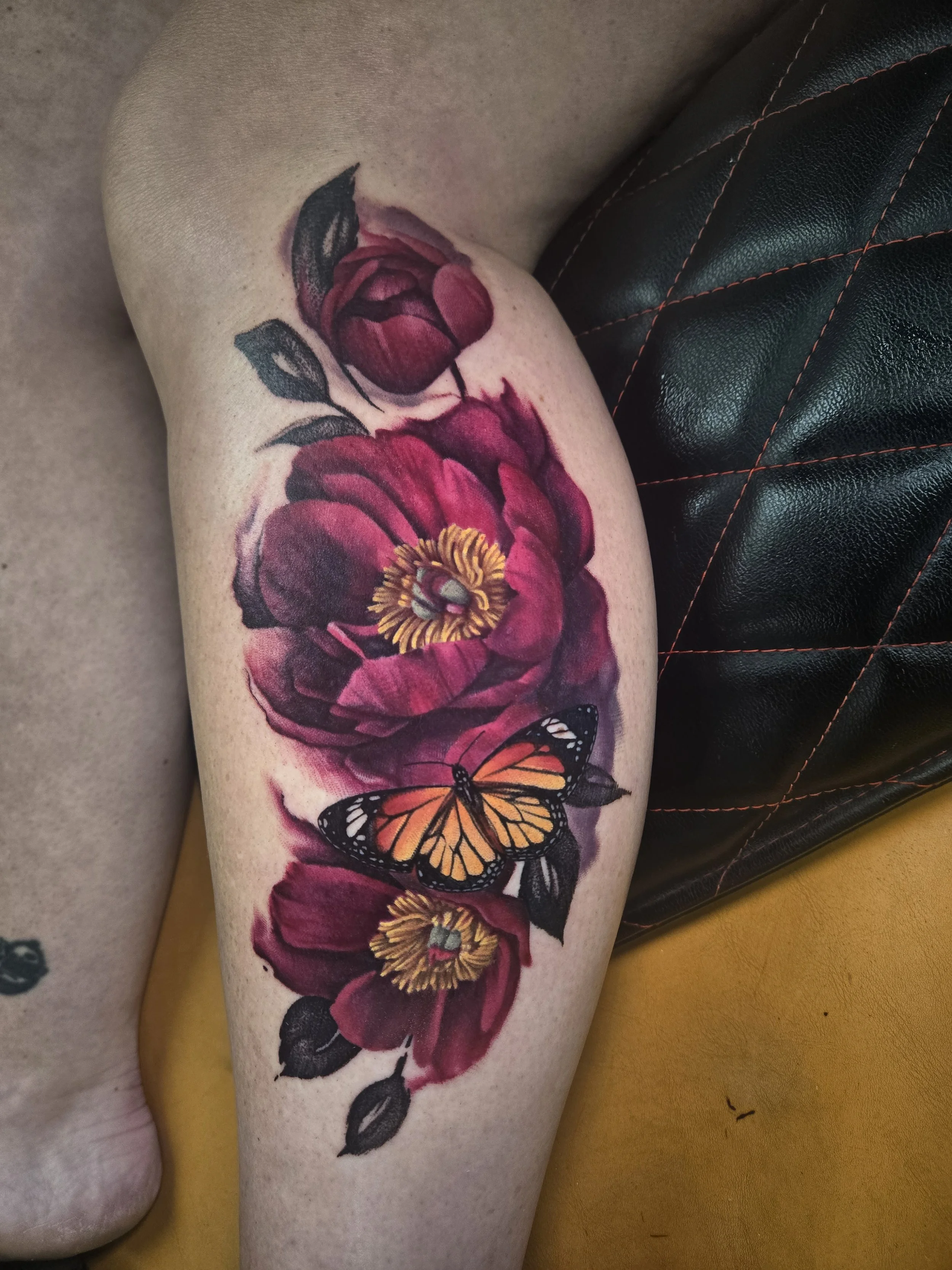 A tattoo of pink and red flowers with green leaves and orange butterflies on a person's calf
