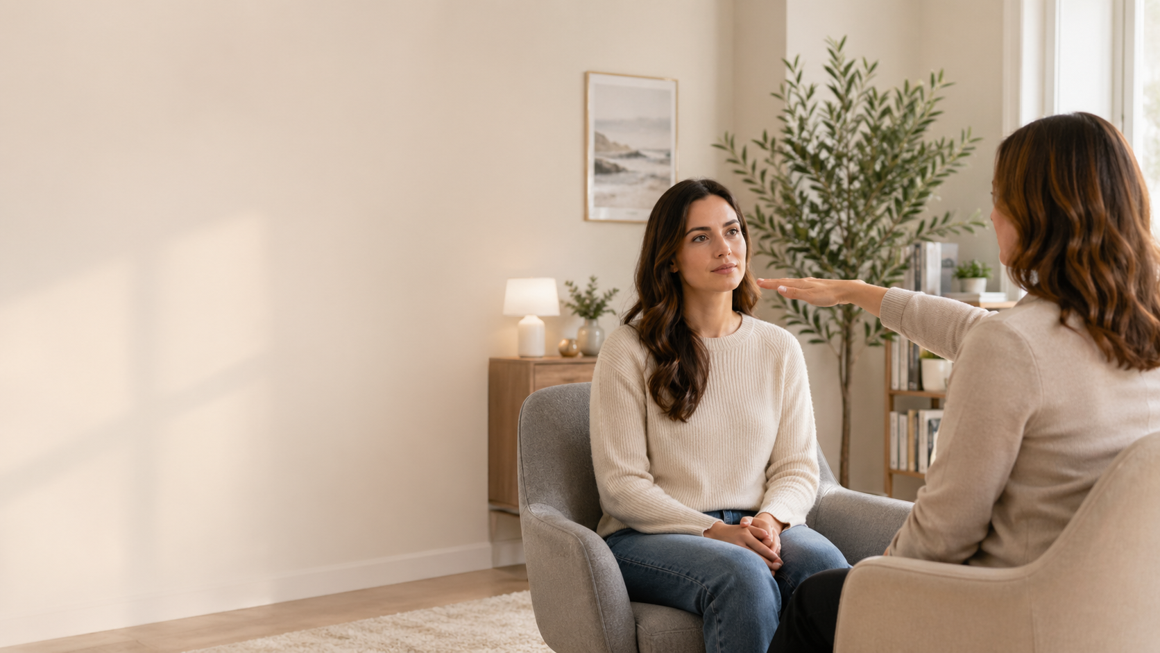 A woman sitting in a therapy session with a therapist in a cozy, well-lit living room. The therapist is holding her hand in front of the woman administering eye movements for EMDR therapy.