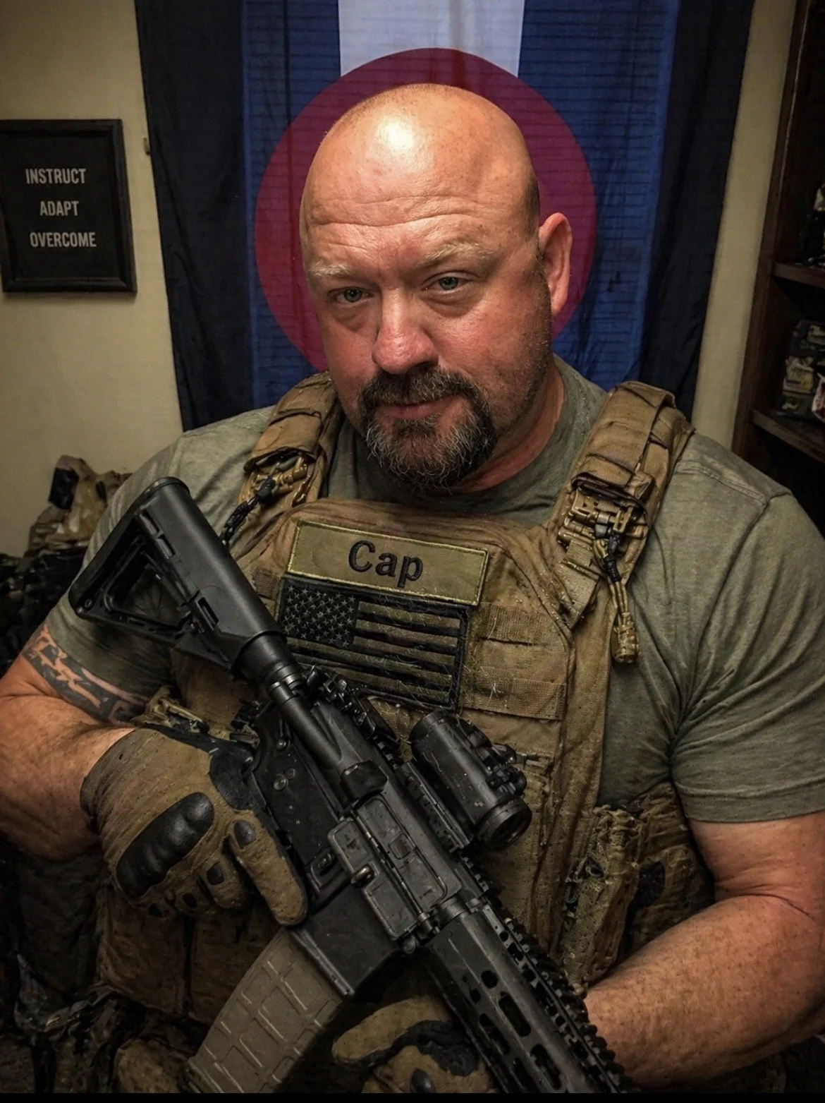 A man dressed in military-style gear, wearing a tactical vest with a name patch labeled 'Cap' and holding a rifle, standing indoors with a background that includes a flag with a red circle and a black background.