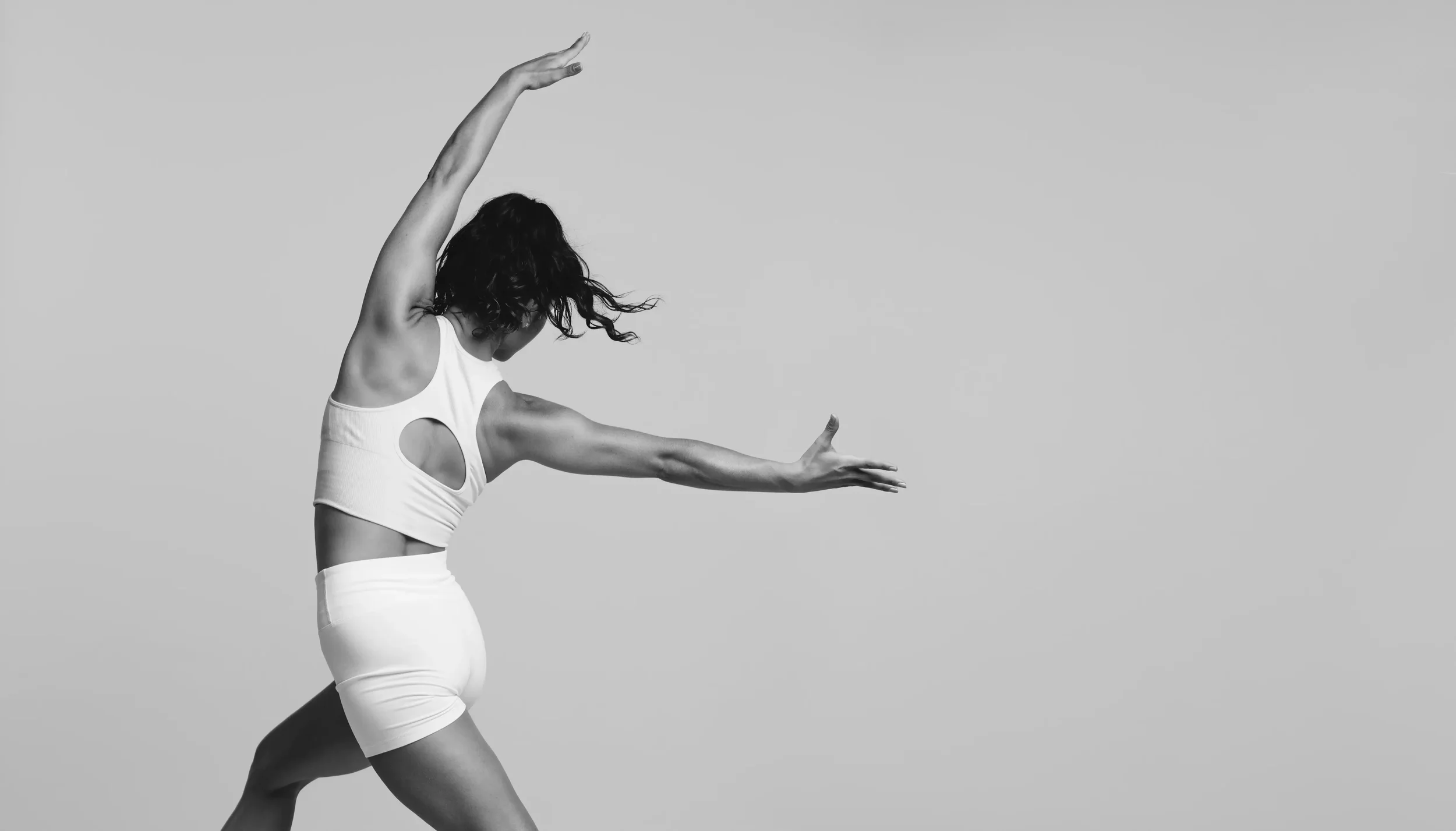 A woman in athletic wear is stretching her arm and leg against a plain background.