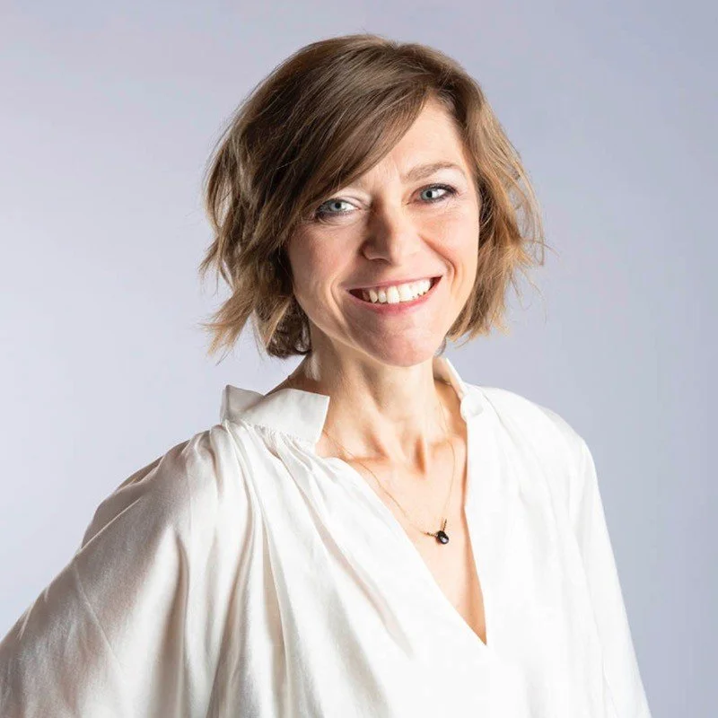 A smiling woman with short, wavy, light brown hair, wearing a white blouse and a small black necklace against a plain background.