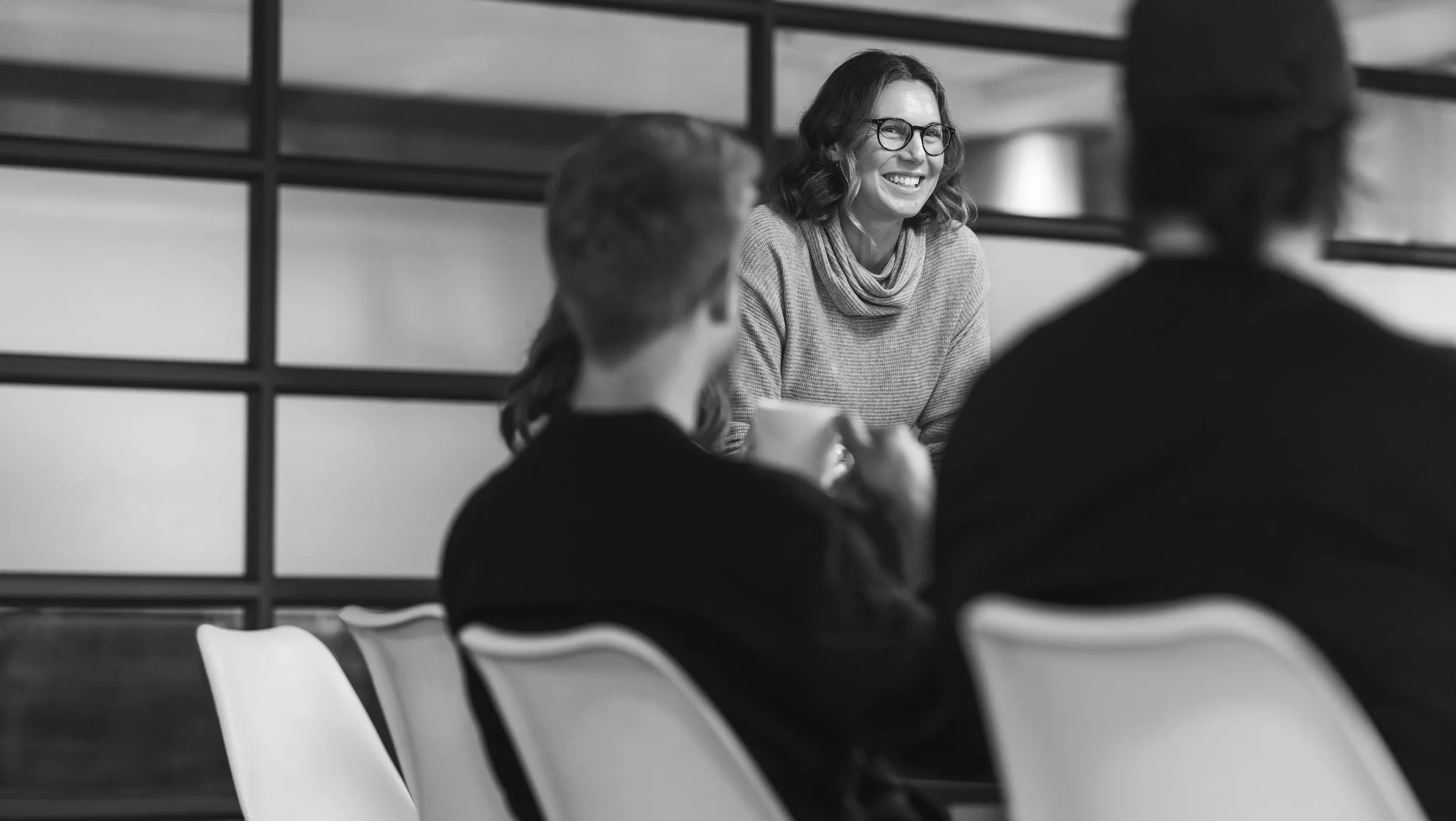 A woman with glasses smiling and talking in a meeting with two other women in a modern office setting, black and white photo.