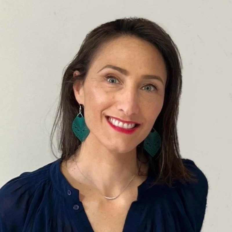 A woman with shoulder-length brown hair smiling and wearing teal earrings and a navy blouse, standing against a plain light-colored wall.
