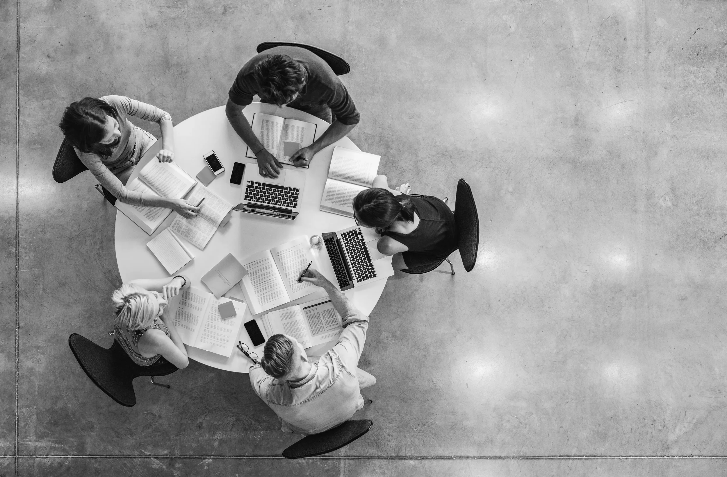 Overhead black and white photo of six people sitting around a round table with open books, laptops, and electronic devices, engaged in studying or discussion in a modern indoor space.