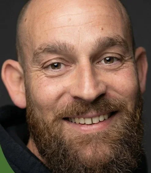 Close-up of a smiling man with a beard and light-colored eyes against a dark background.