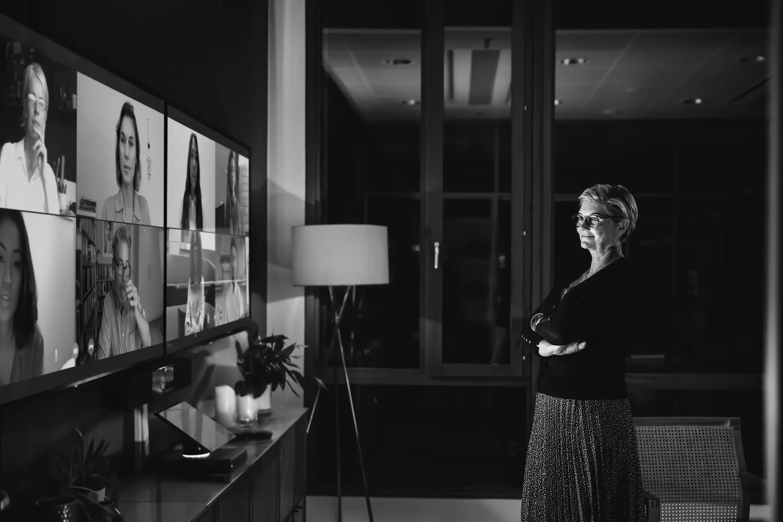 A woman with glasses and short hair stands with crossed arms in a dimly lit room, looking at a wall of screens showing video call participants.