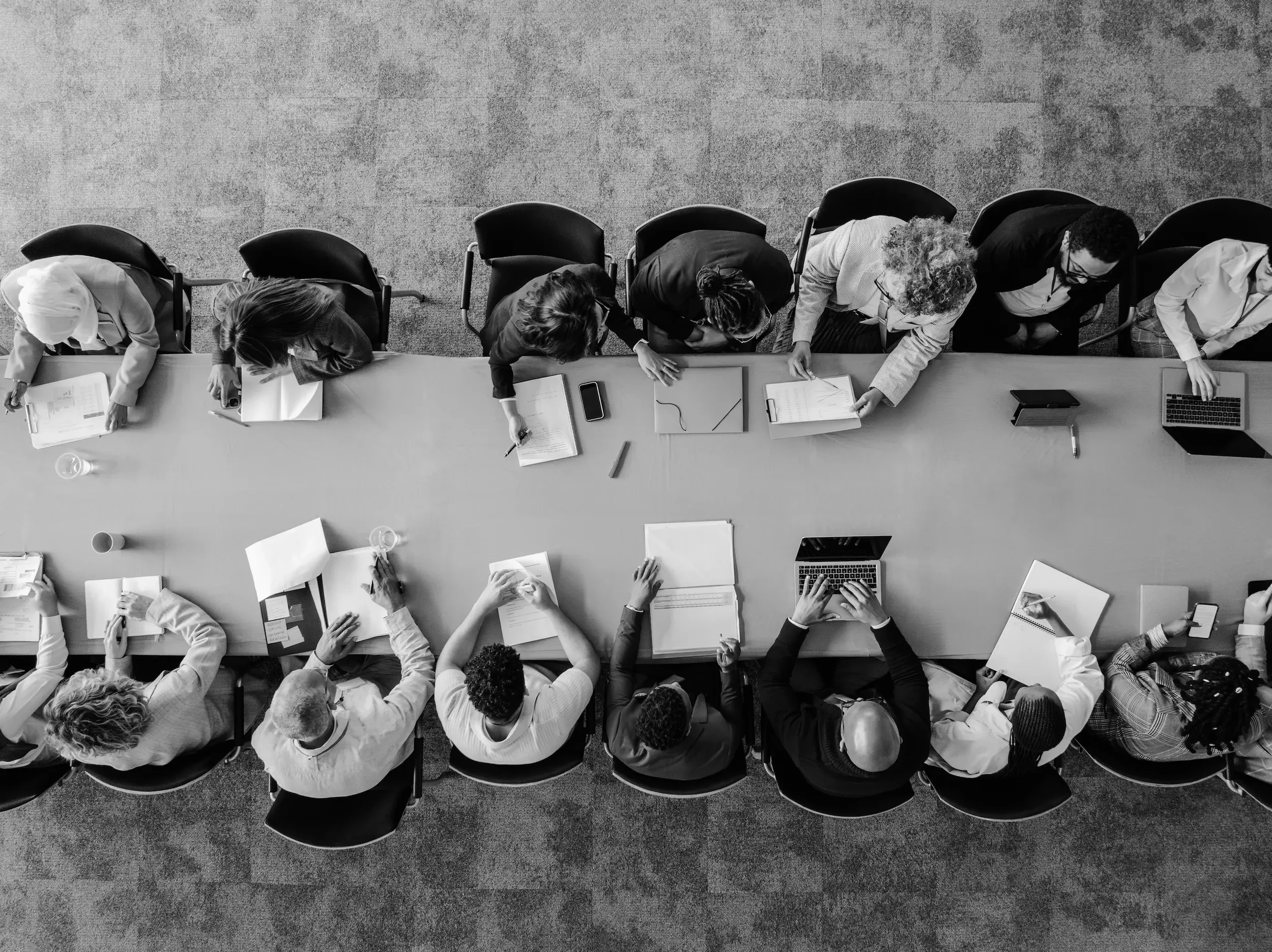Top-down black and white photo of a group of people sitting around a long table, working on documents, using laptops, and taking notes.