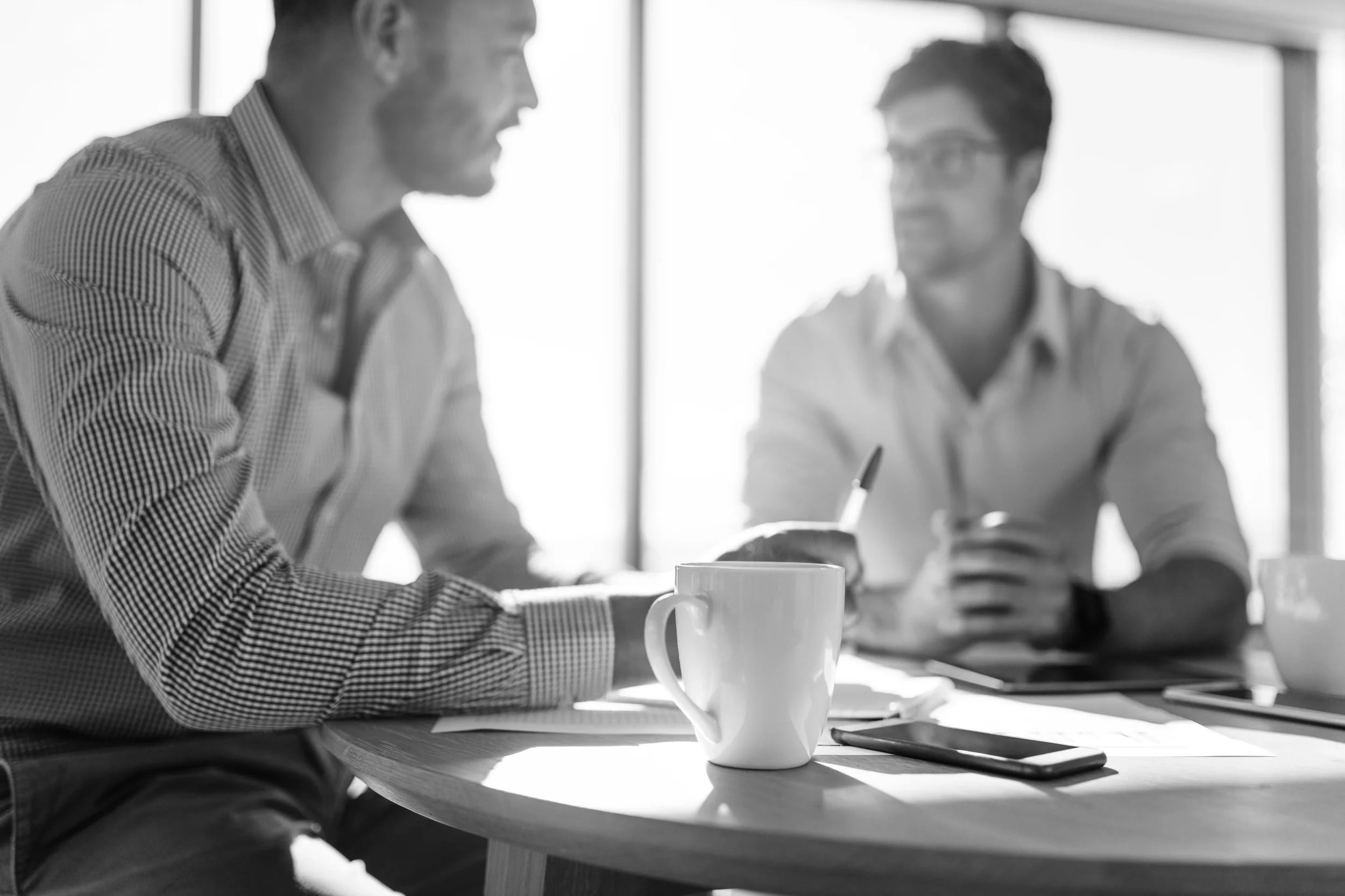 Two men are sitting at a round table in a meeting room, engaged in conversation. One man in the foreground is wearing a checkered shirt, and a large cup is on the table in front of him. The other man, in the background, is wearing glasses and a light-colored shirt, holding a pen, with papers and a smartphone on the table.