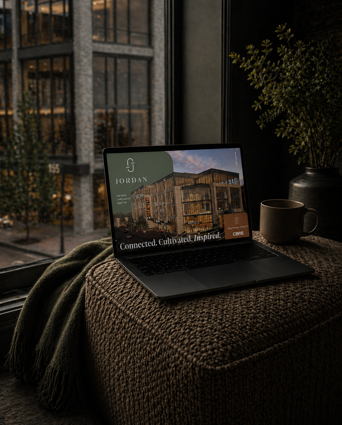 A laptop on a textured ottoman shows a real estate advertisement for Jordan with a building and the tagline "Connected. Cultivated. Inspired." next to a mug and a potted plant, with city buildings and trees visible outside a window.