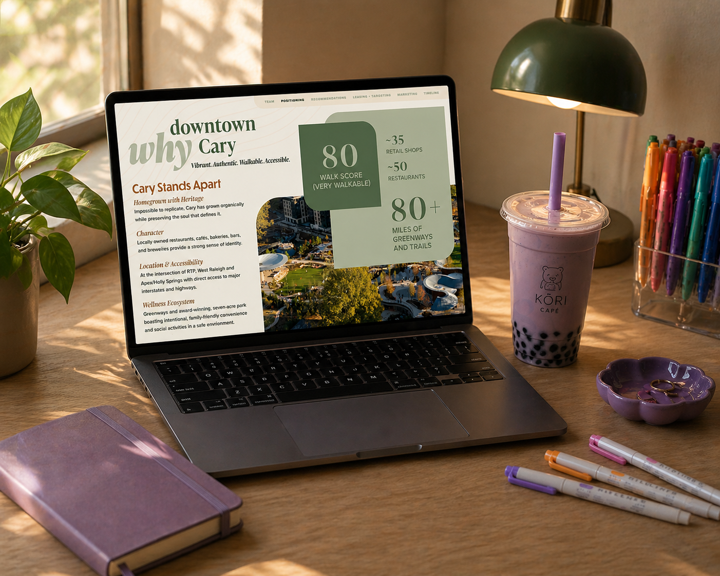 A desk workspace with an open laptop displaying a website, a purple notebook with an elastic band, a bubble tea drink with a purple straw, a purple flower-shaped dish with rings, a set of colorful markers, and a small green plant, positioned next to a window with natural light.