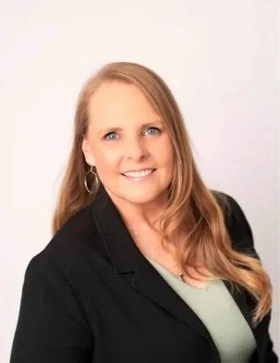 A woman with long blonde hair wearing a black blazer and light-colored top, smiling at the camera against a plain white background.